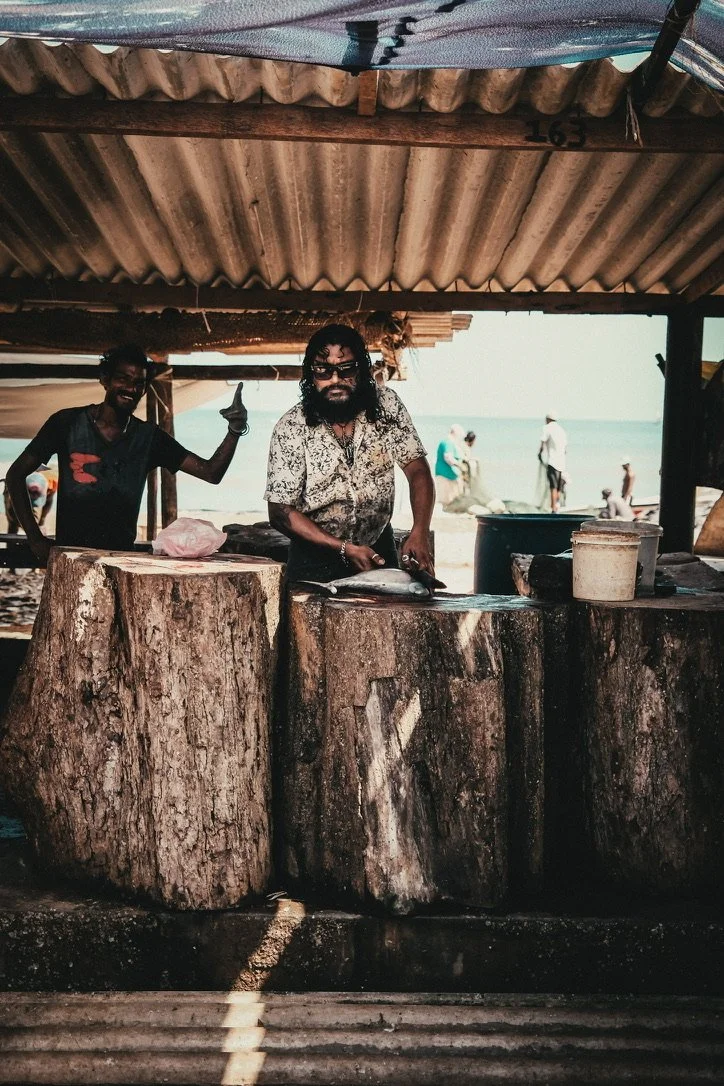 Men preparing fish on wooden logs at a beachside stall with the ocean and beachgoers in the background.