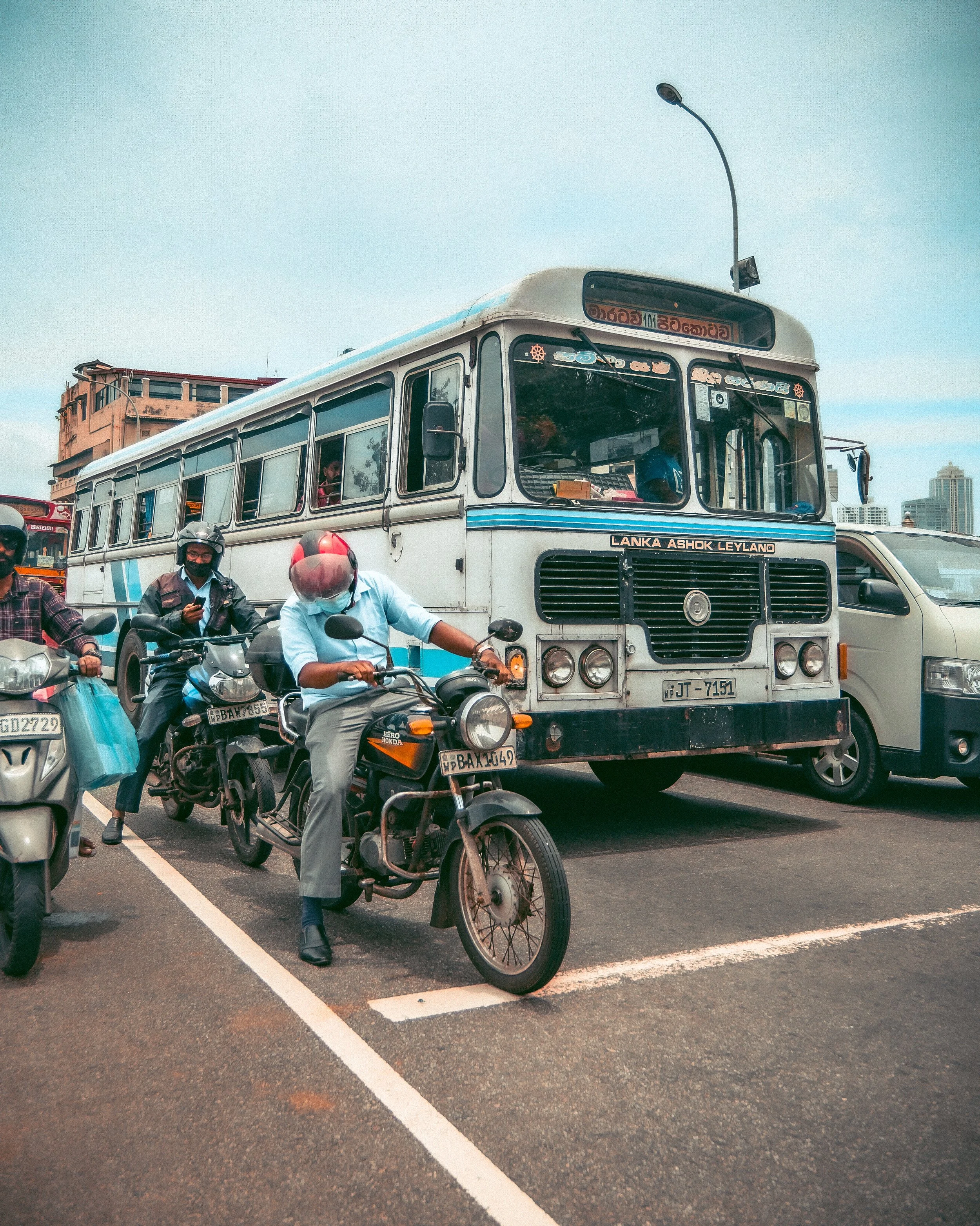 Street scene with motorcycles and a bus in traffic, with buildings and a cloudy sky in the background.