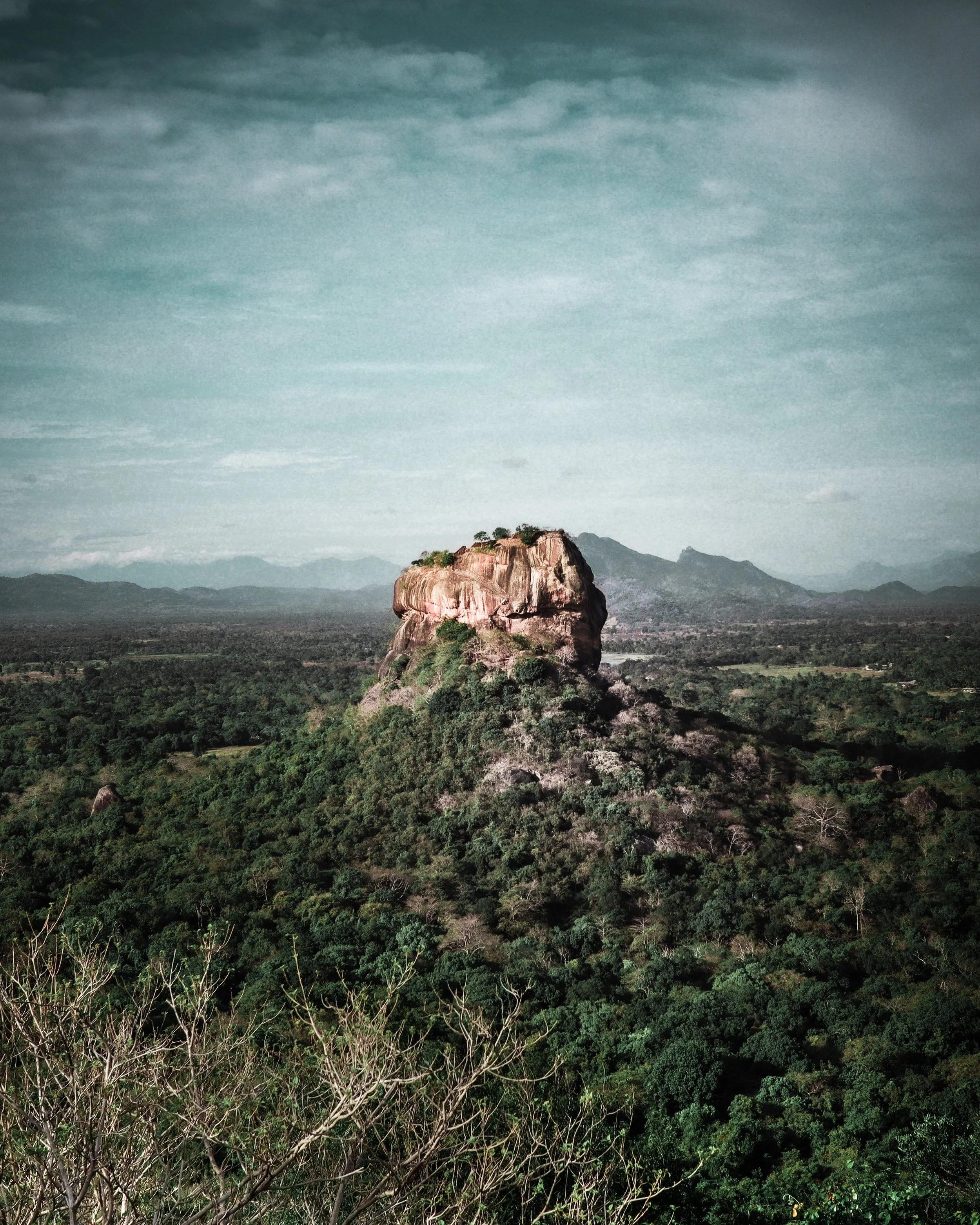 Large rock formation surrounded by green forested hills and distant mountains under cloudy sky