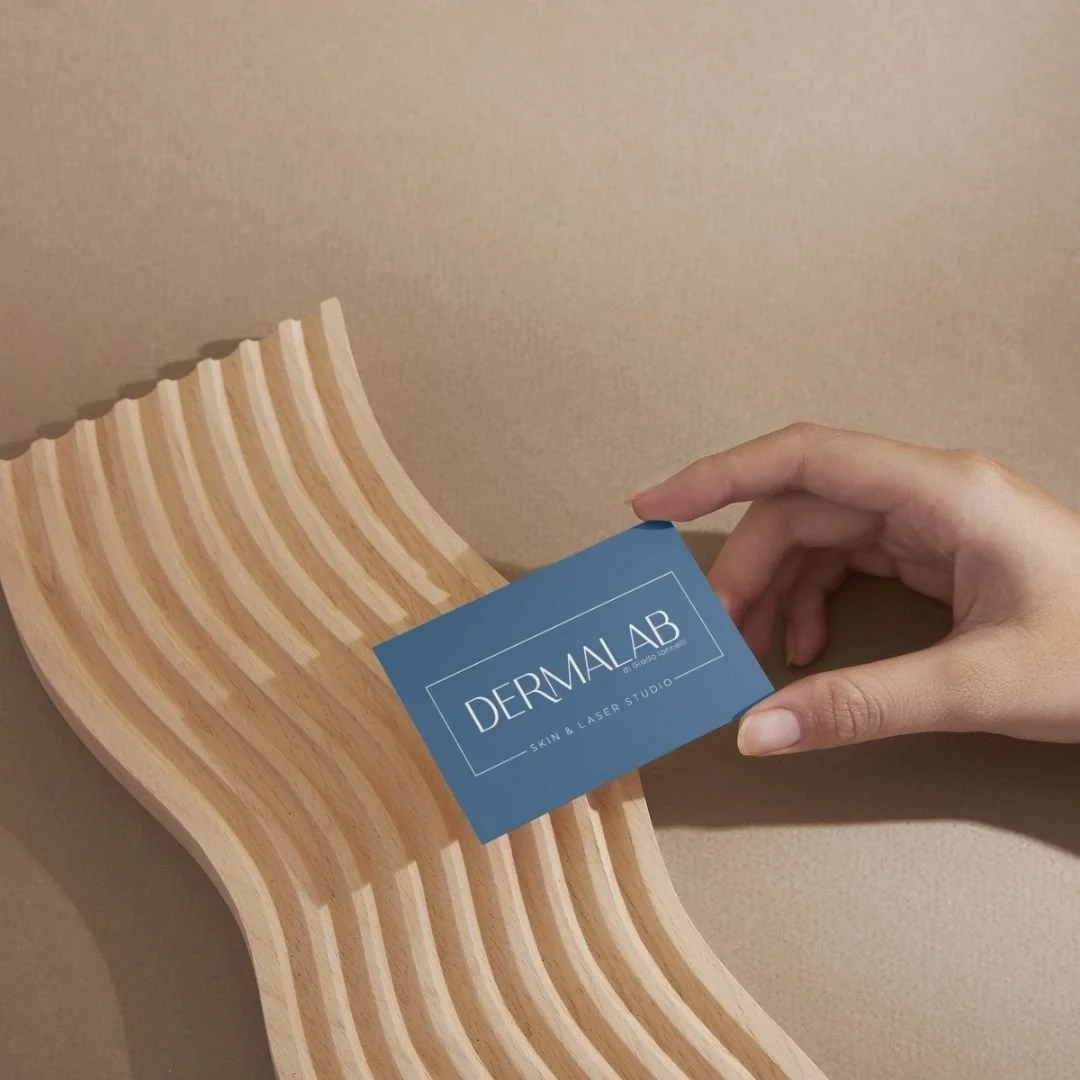 Mano che tiene un biglietto da visita con scritto 'DERMALAB SKIN & LASER STUDIO' di fronte a una sedia di legno con design curvo.