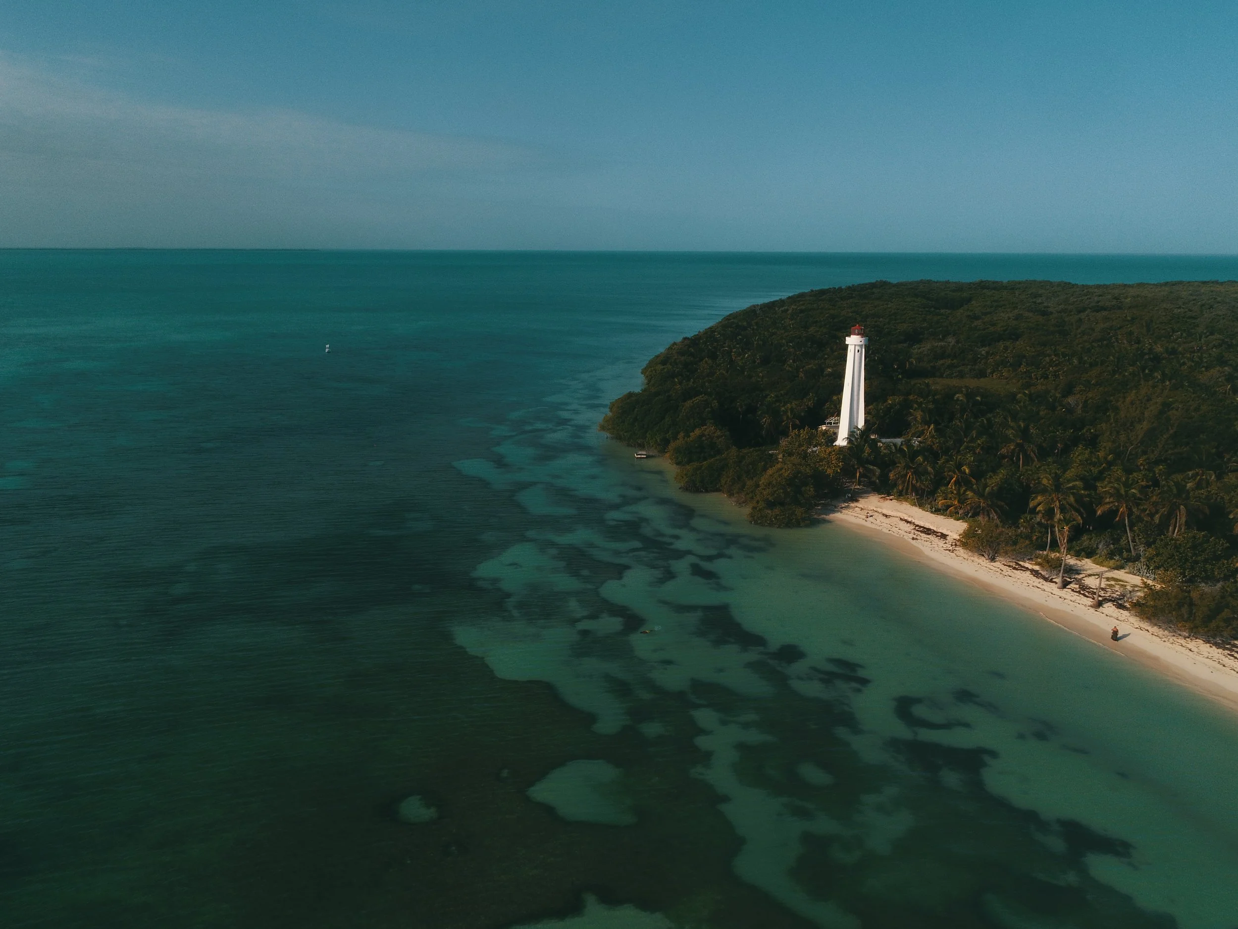 Aerial view of a lighthouse on a green, tree-covered island with a beach, turquoise waters, and the open ocean in the background.