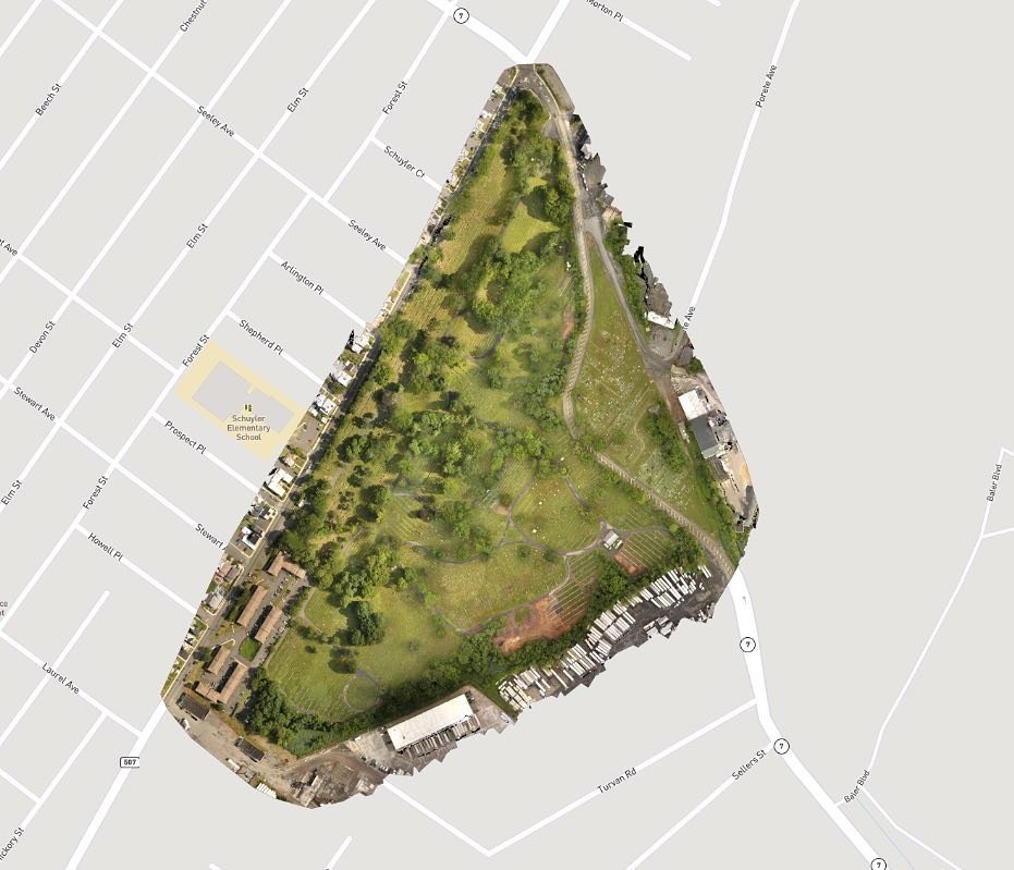 A satellite image of a triangular park surrounded by roads, with trees, open grassy areas, pathways, and some buildings.