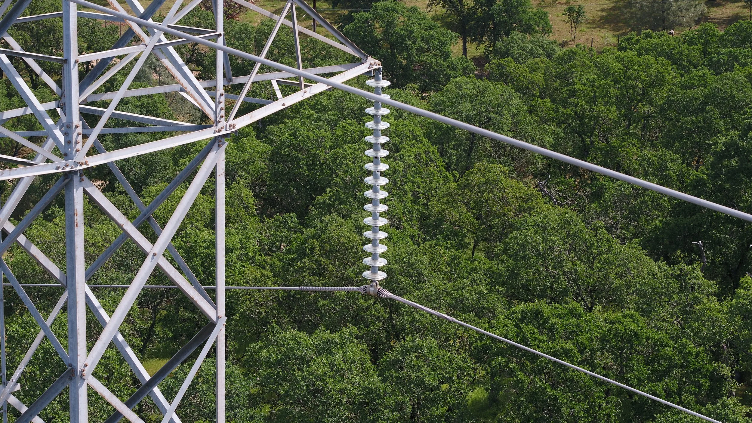 Close-up of a metal electrical tower with insulators and power lines extending into a green forested landscape.