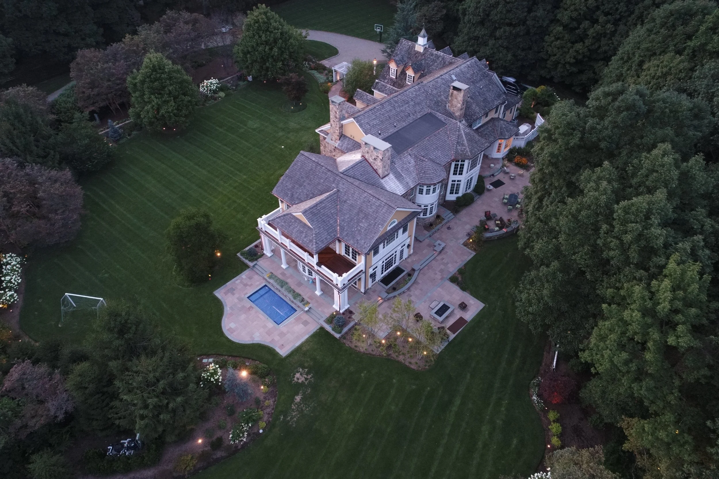 Aerial view of a large, elegant house with multiple chimneys and a backyard with a lawn, a small swimming pool, a sports court, and outdoor seating surrounded by trees.