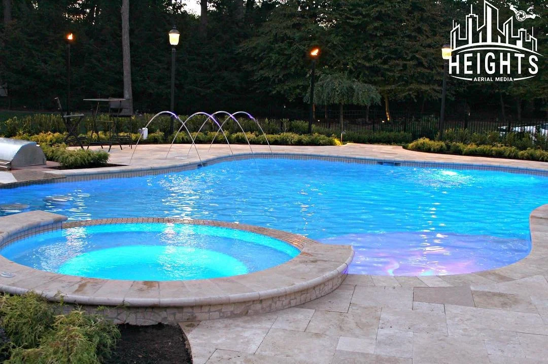 A swimming pool and hot tub at dusk with illuminated water features, surrounded by a tiled deck, outdoor furniture, and lush greenery with trees in the background.