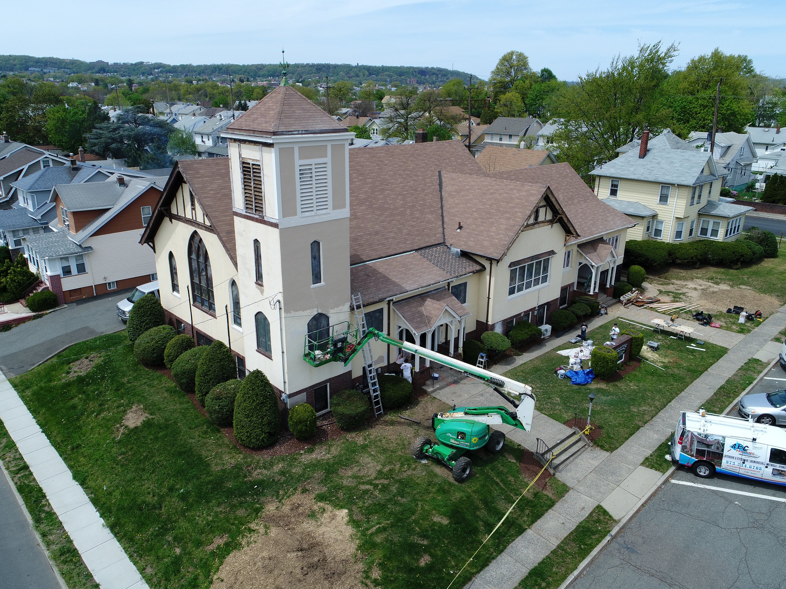 A white church with a brown roof and a tall steeple under renovation, surrounded by a well-manicured lawn and bushes, with workmen and equipment including a lift, ladders, and a maintenance van nearby.