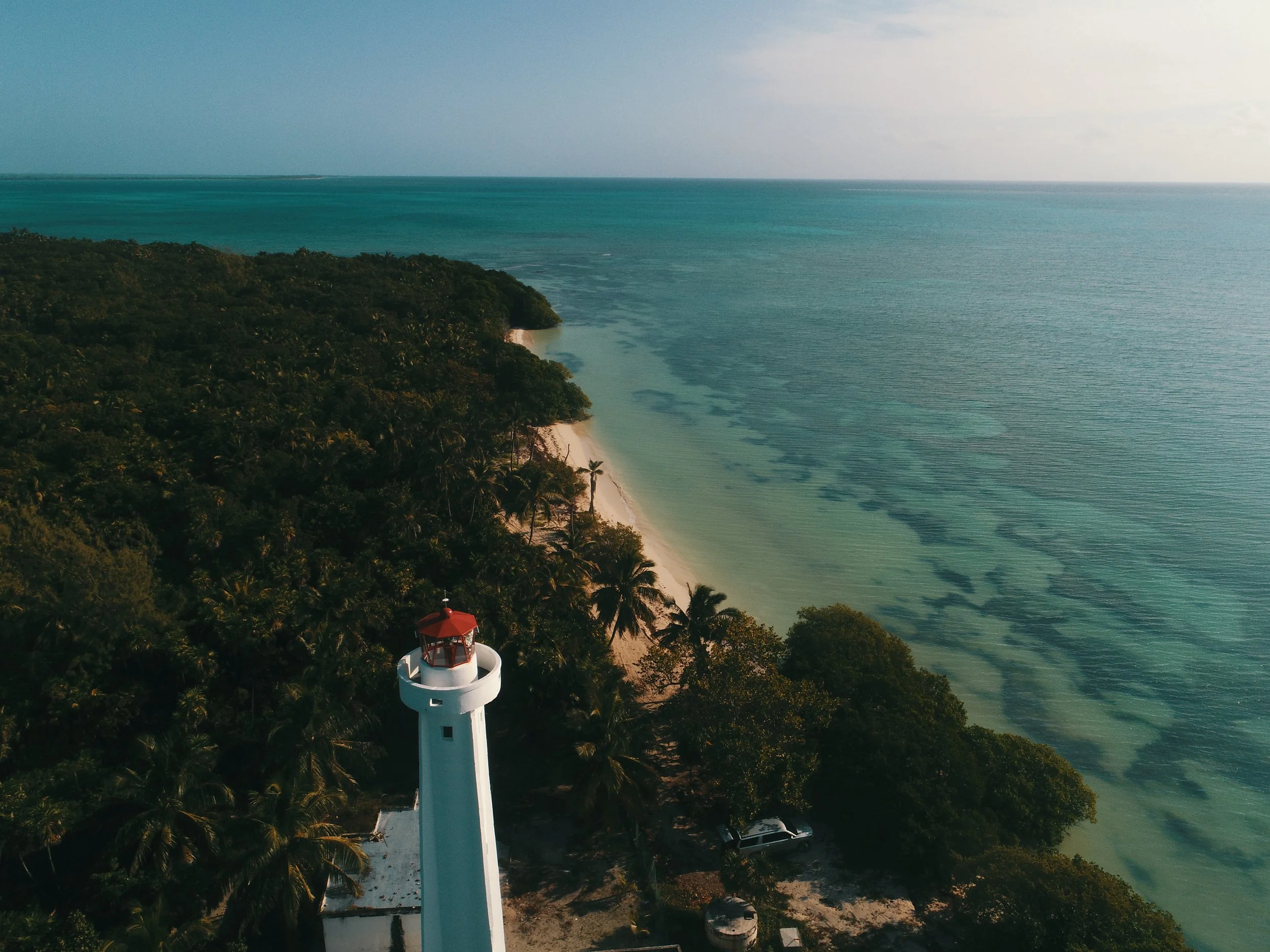 An aerial view of a white lighthouse with a red top standing among lush green trees along a sandy coastline with turquoise water and gentle waves, with a distant horizon and cloudy sky.