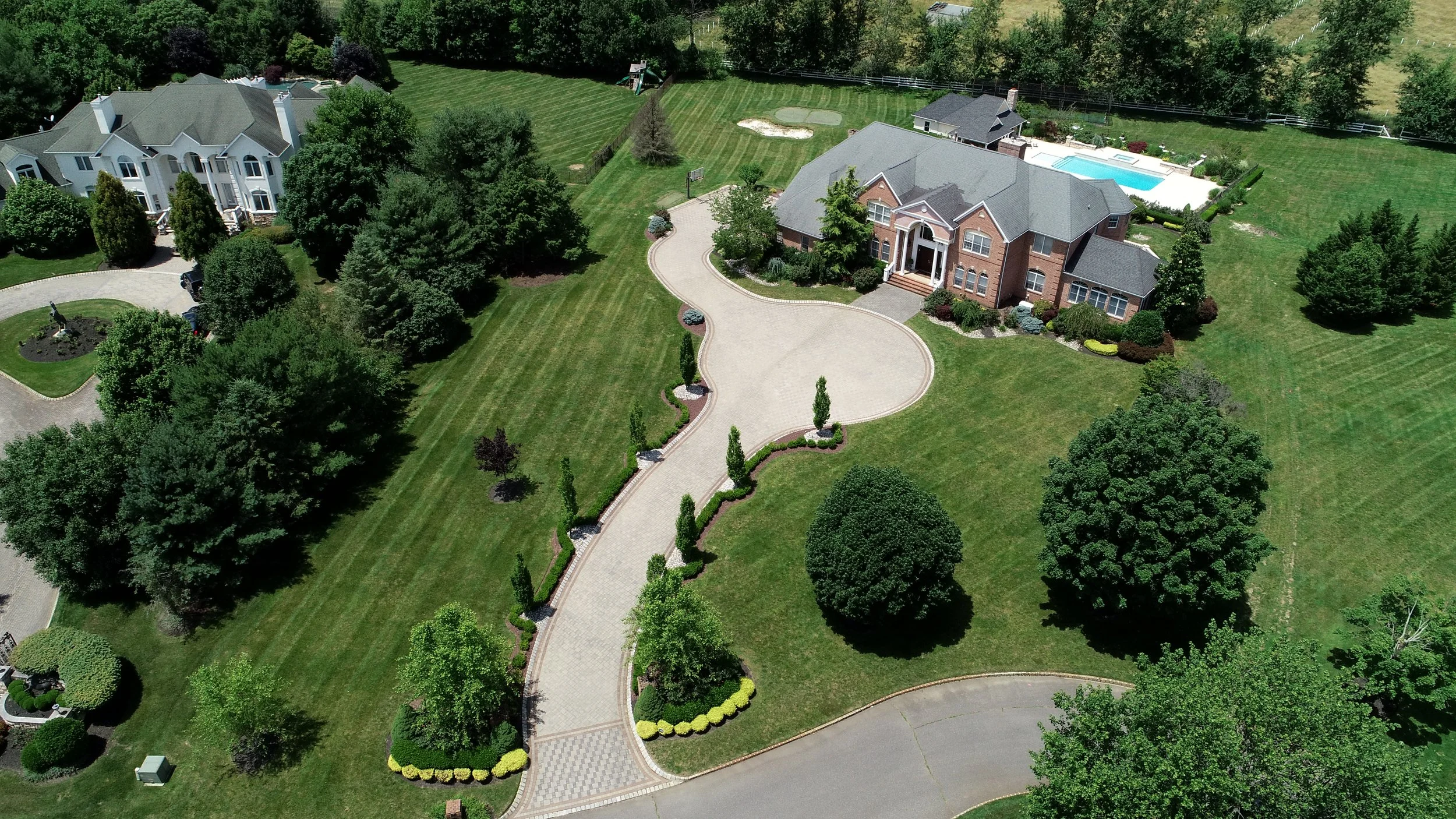 An aerial view of a large brick house with a grey roof, surrounded by a well-manicured lawn with trees, bushes, and a paved entrance driveway. There is a swimming pool in the backyard and a smaller building nearby. The property is enclosed by a fence with surrounding greenery.