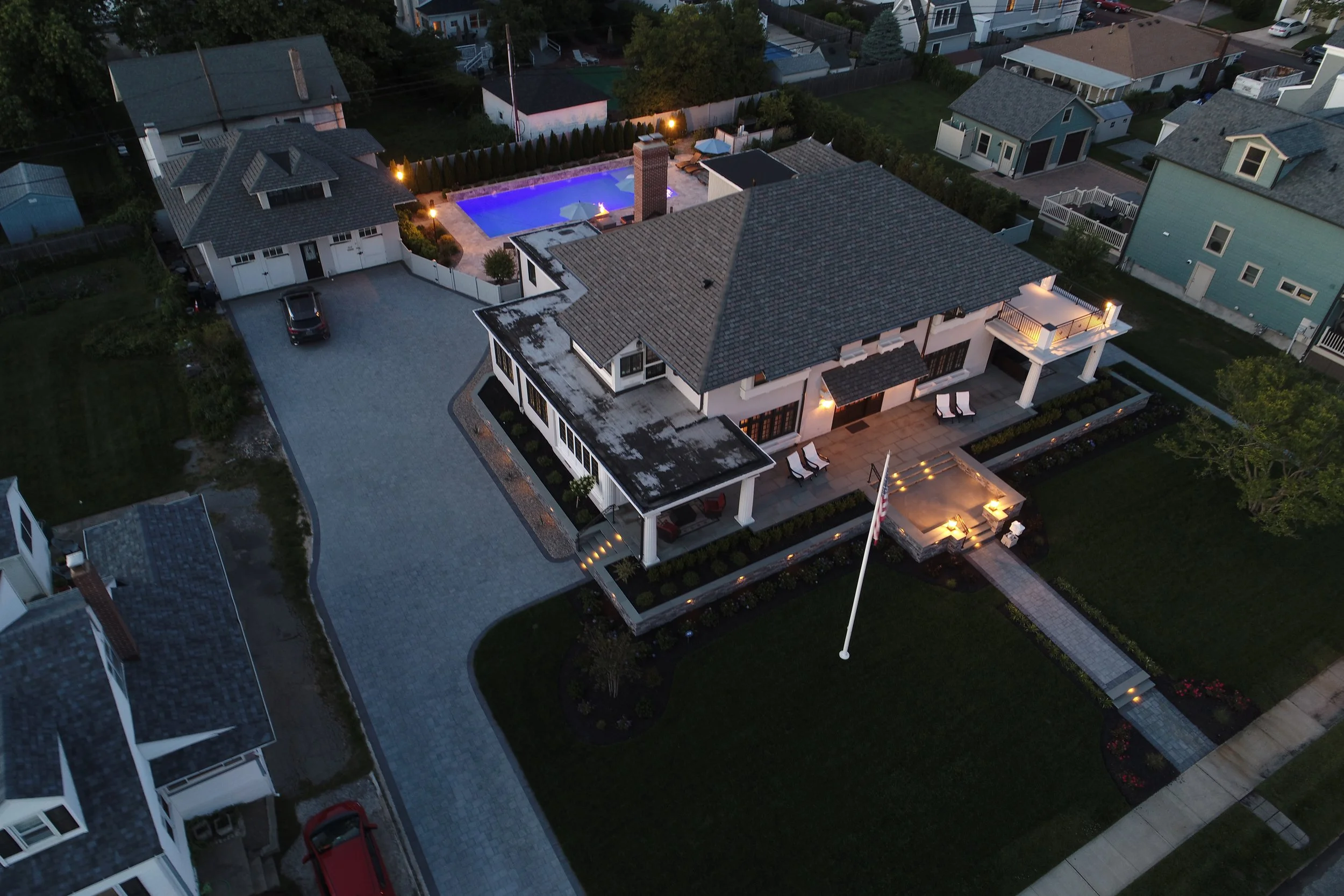 Aerial view of a large house with a patio, driveway, and a backyard swimming pool illuminated at night. Neighboring houses are visible around.