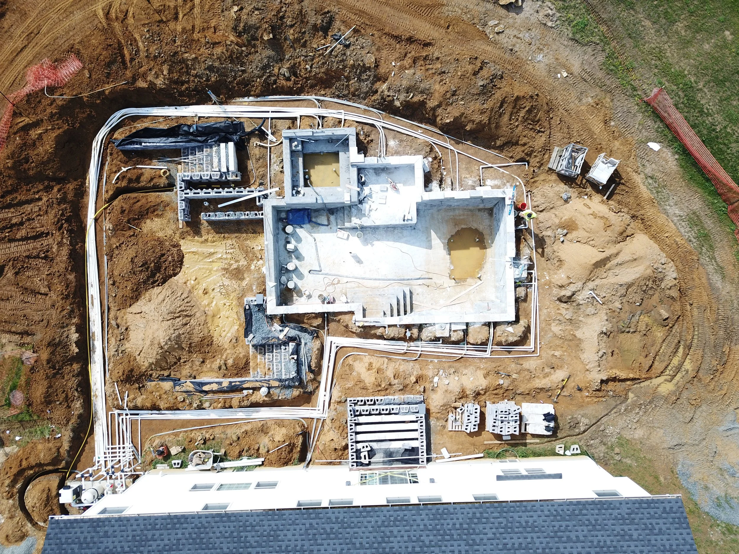 An aerial view of a house under construction with exposed foundation, building materials, and construction workers in a dirt lot, surrounded by a yard with gravel and construction fencing.