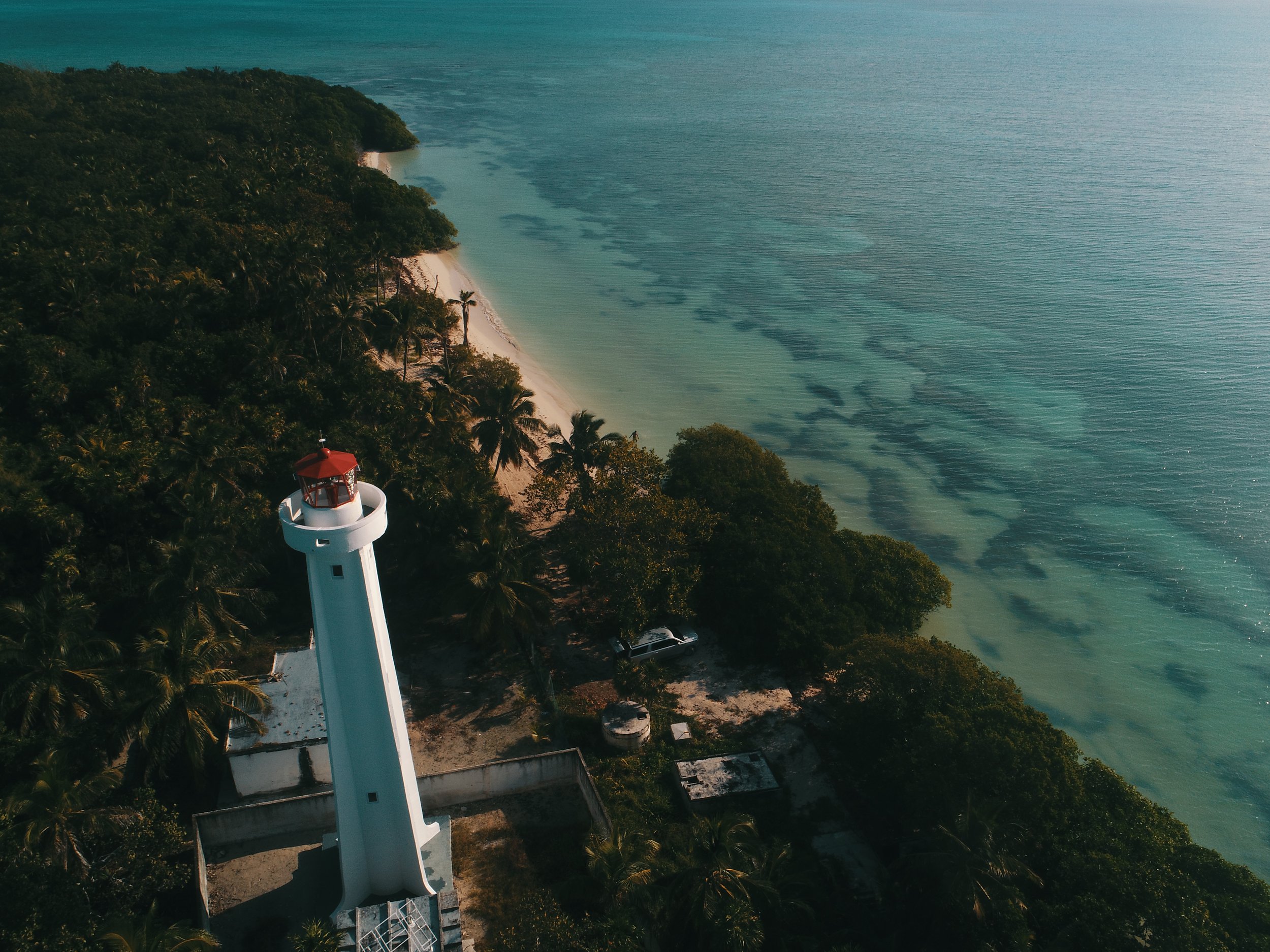 Aerial view of a lighthouse on a tropical beach with palm trees and clear blue water.