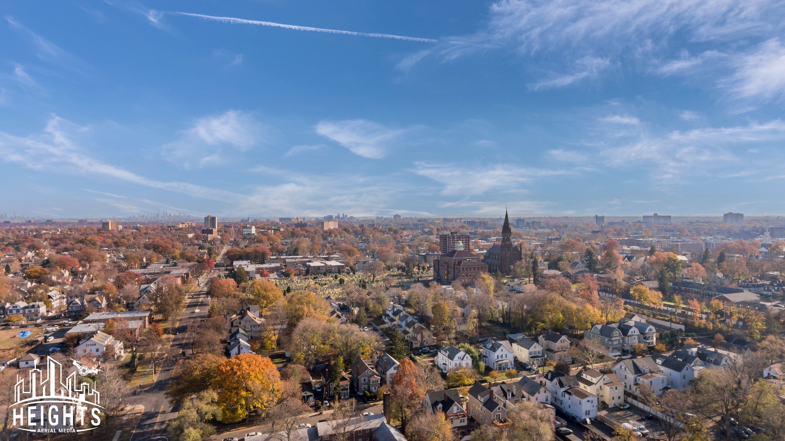 Aerial view of a city with a mix of houses and taller buildings under a partly cloudy blue sky, with trees showing fall colors.