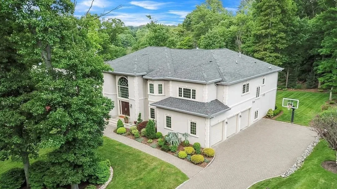 Large white house with a dark gray roof, surrounded by green trees and a well-maintained lawn. A concrete driveway leads to a three-car garage, and there is a small basketball hoop on the side.