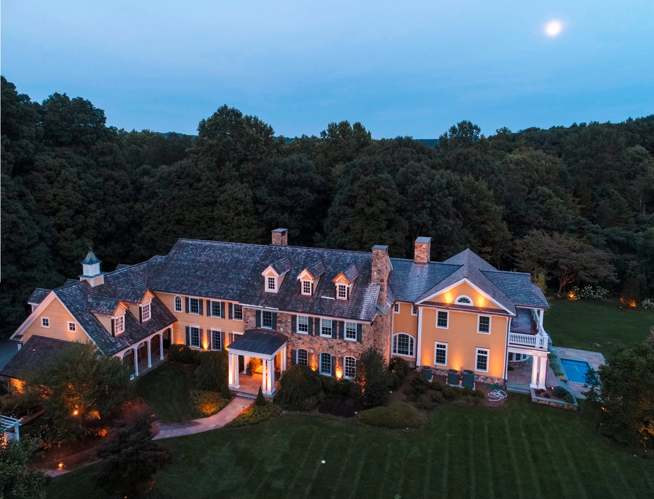 A large yellow mansion with multiple chimneys, a covered porch, and a swimming pool, set in a lush green backyard with trees in the background, at dusk.