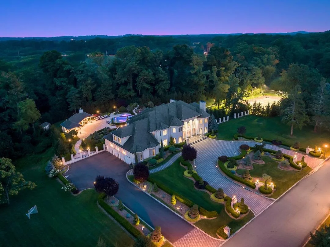 A large, elegant house with a driveway, manicured lawn, and surrounding trees, illuminated at night with outdoor lighting.