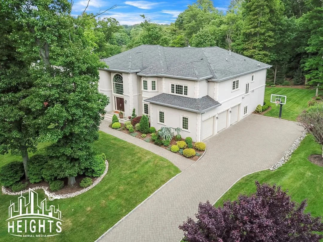 A large two-story house with a gray roof, surrounded by lush green trees and landscaped yard. There is a paved driveway leading to a three-car garage and a small basketball hoop nearby.