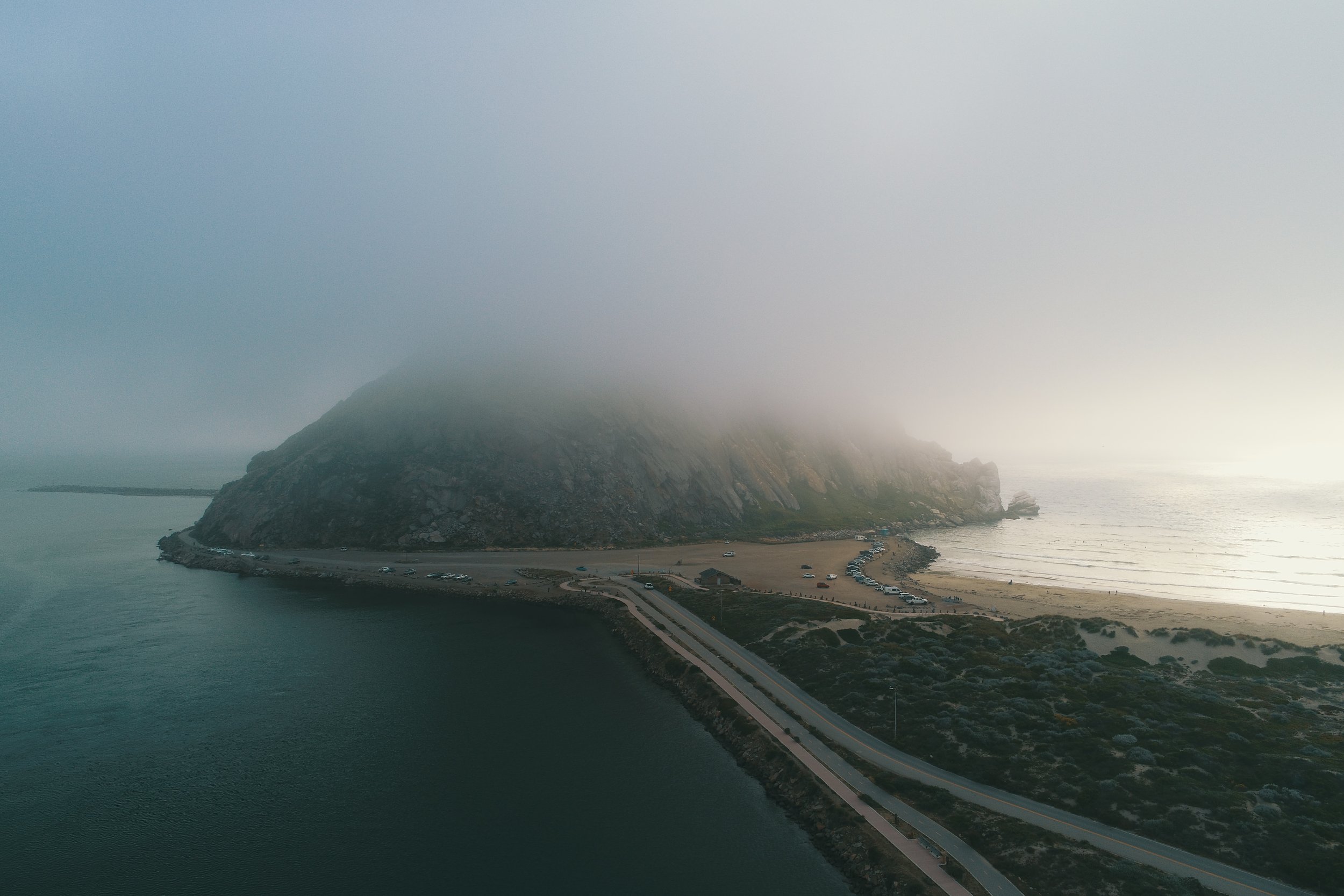 A foggy coastal landscape featuring a large rocky hill or mountain with mist covering its top, situated near a sandy beach and calm water. There is a winding road along the shoreline with cars parked nearby.