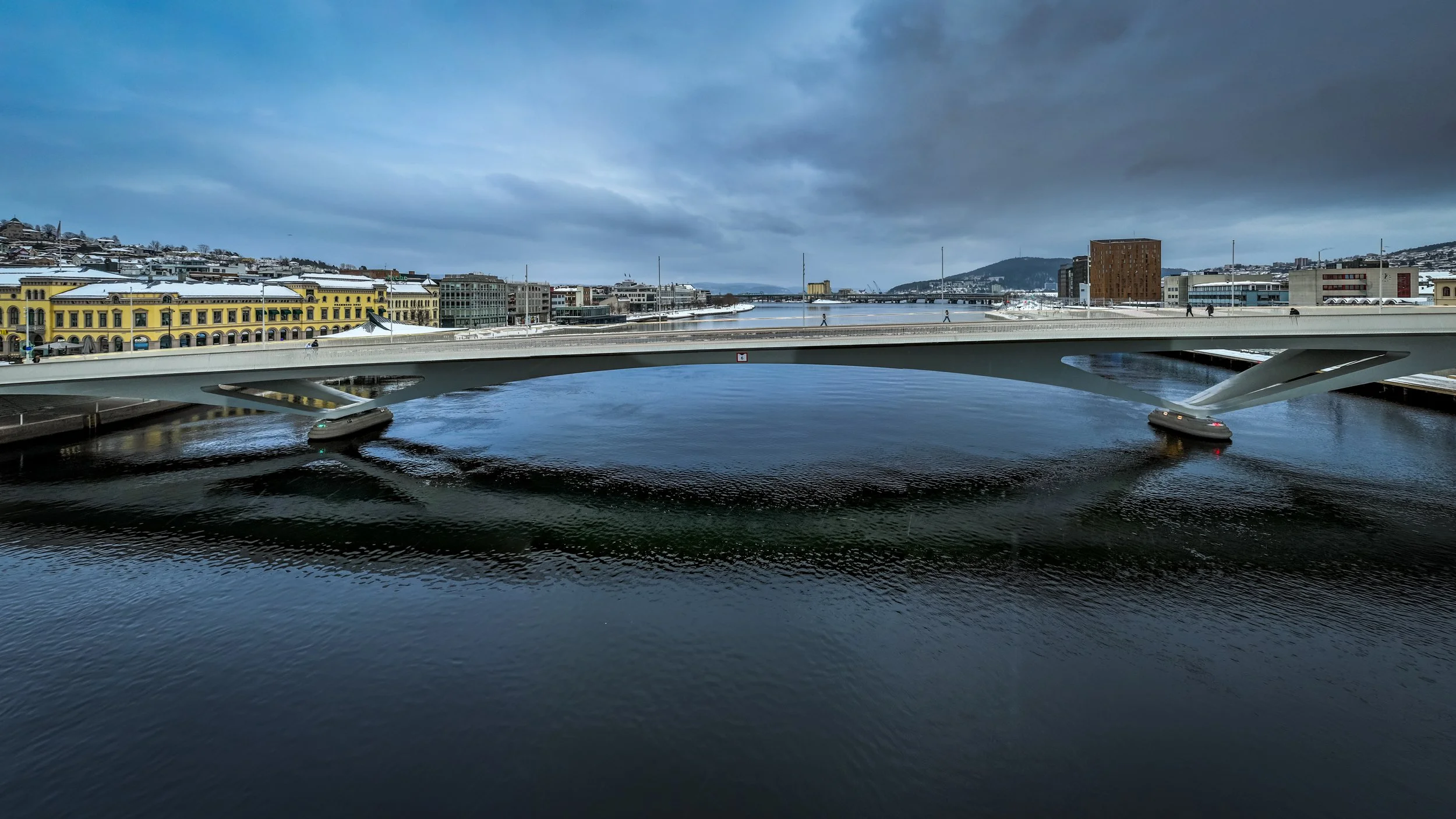 A modern white bridge over a river in a city with colorful historic buildings and a snowy hillside in the background under a cloudy sky.