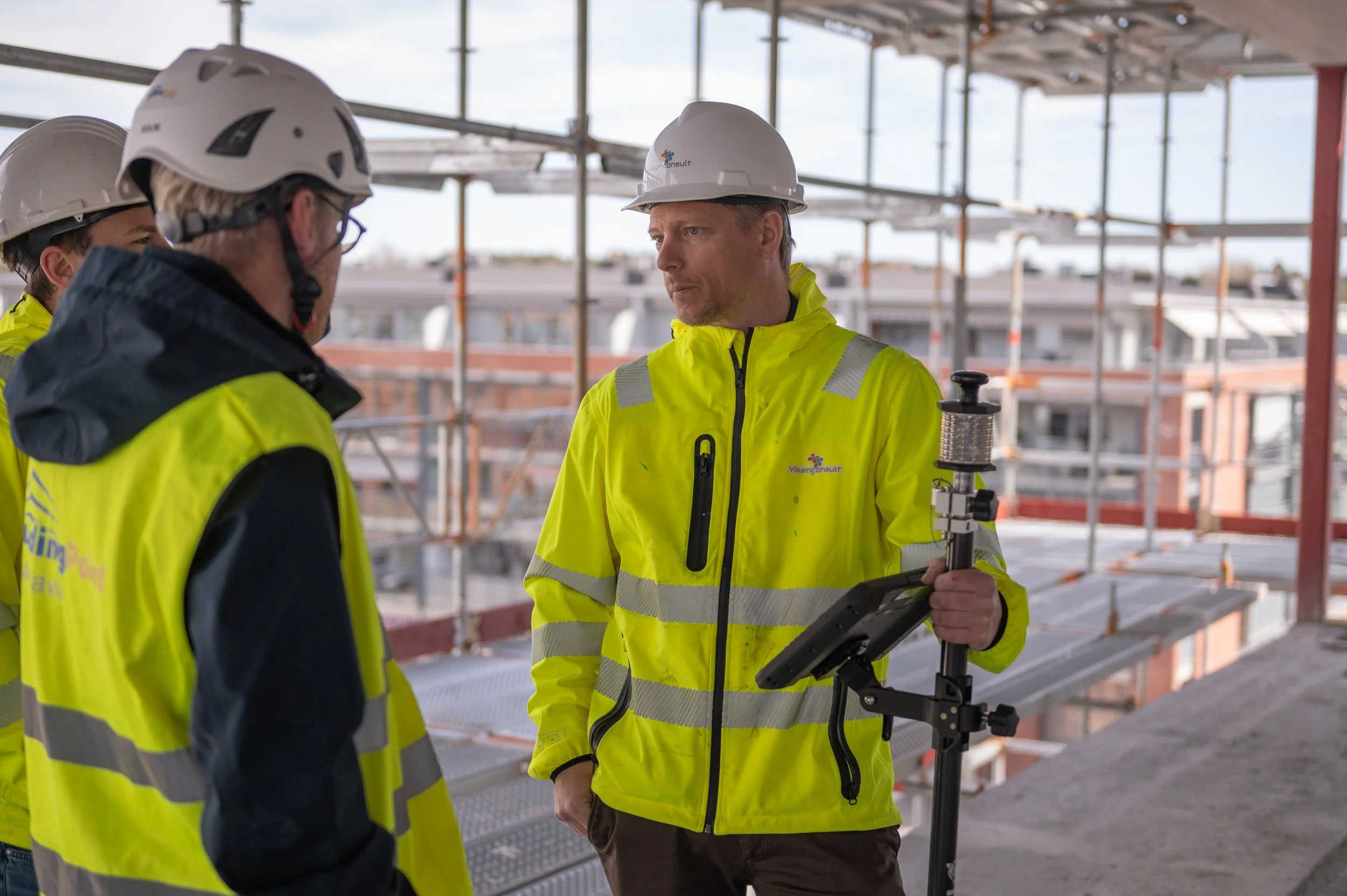 Three construction workers wearing yellow high-visibility jackets and white hard hats on a construction site. One worker holds a tablet device and appears to be explaining something to the others.