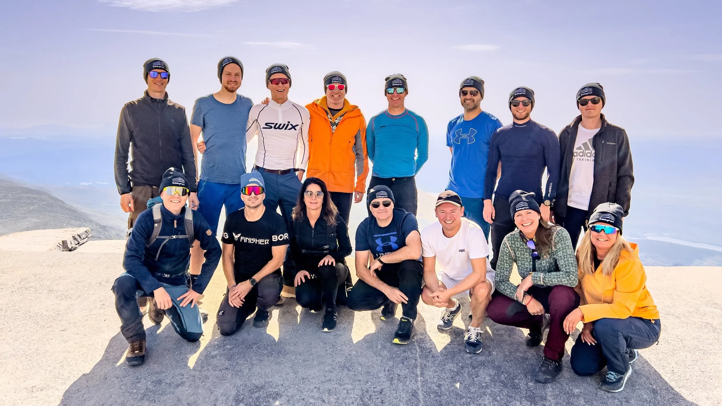 Group of 13 people in outdoor mountain clothing, some wearing sunglasses and hats, posing for a photo at the summit of a mountain with cloudy sky in the background.