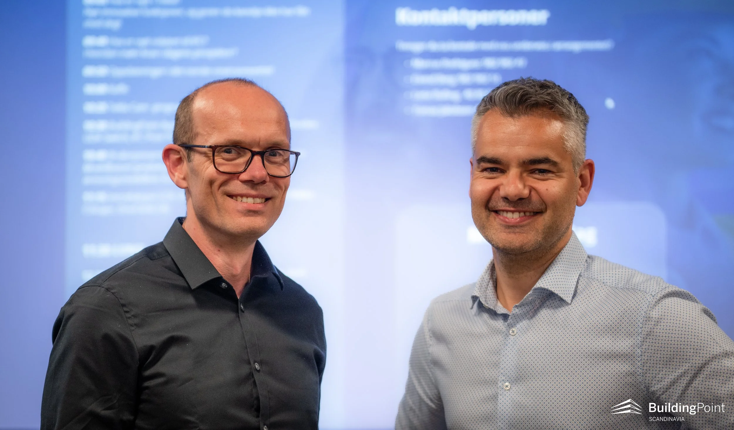 Two men smiling and standing side by side in front of a blue screen with white text. The man on the left is wearing glasses and a black shirt; the man on the right has gray hair and is wearing a light-colored dress shirt. The BuildingPoint Scandinavia logo is visible in the bottom right corner.