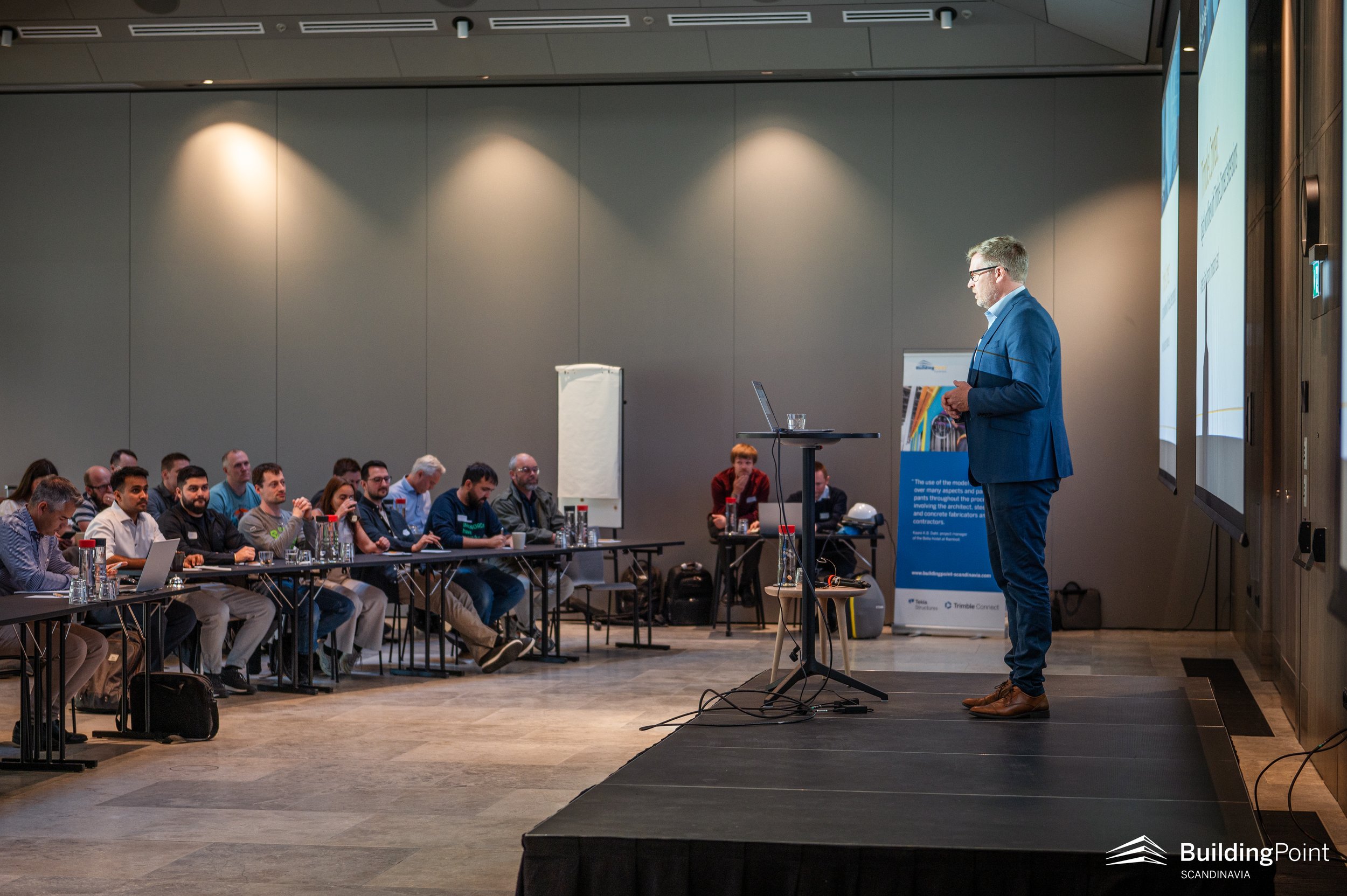 A man in a blue suit giving a presentation to an audience seated at tables in a conference room, with a laptop and a projector screen displaying information.