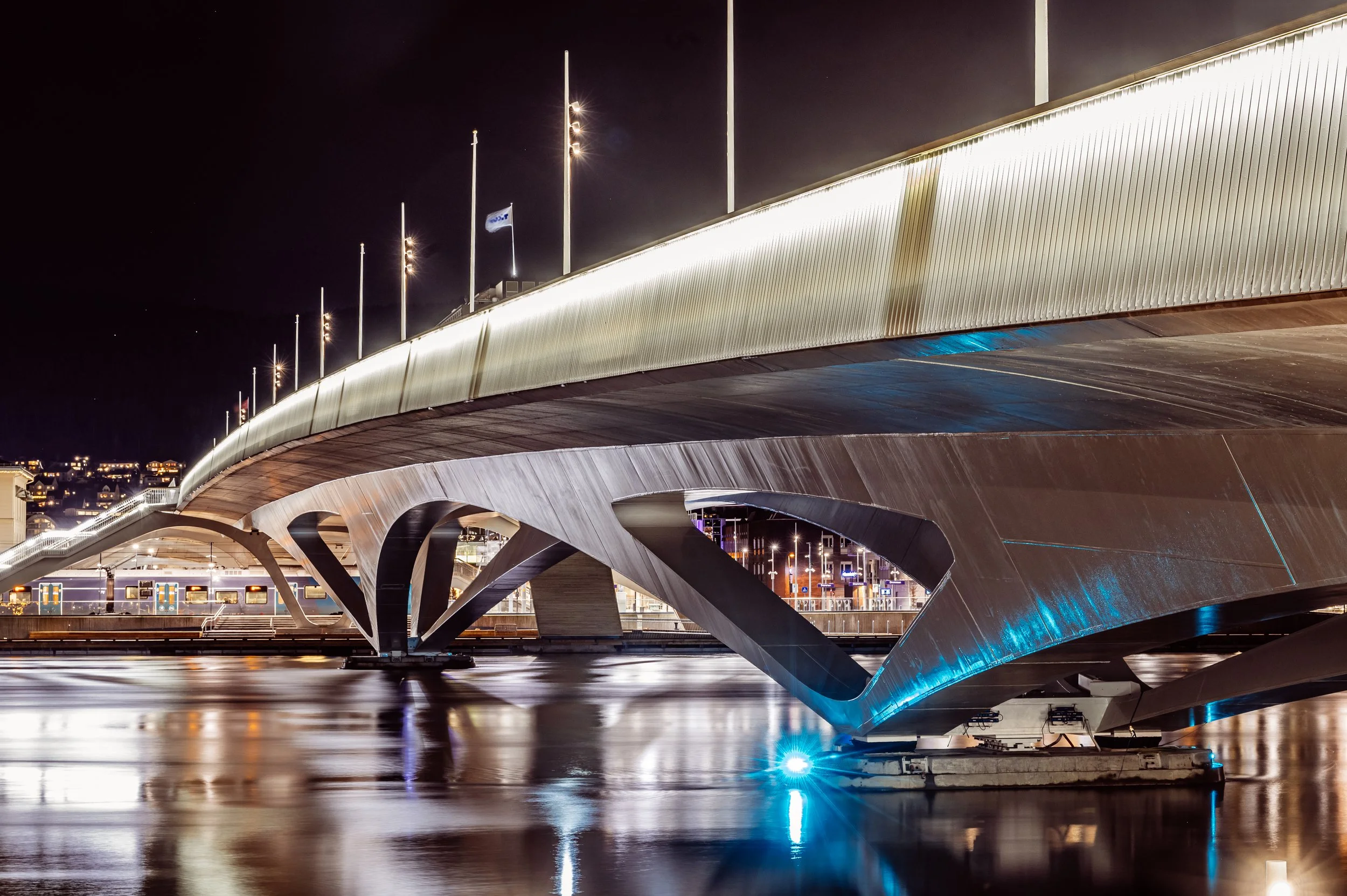 Night view of a modern, illuminated bridge with curved metal supports reflecting in the water below, city lights visible in the background.