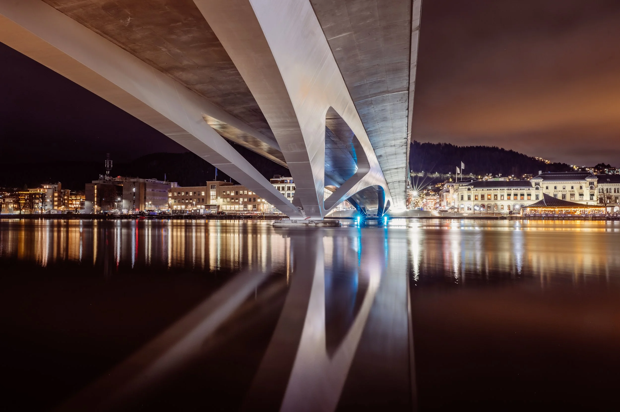 Nighttime view of a modern bridge with illuminated underside, reflecting in water below, with city buildings and hills in the background.