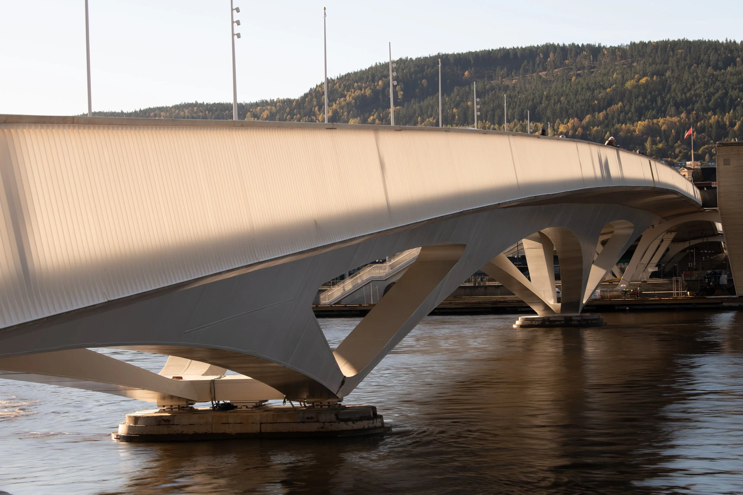 Modern white bridge with curved design spanning over water, with people walking on it and a background of green mountains with trees