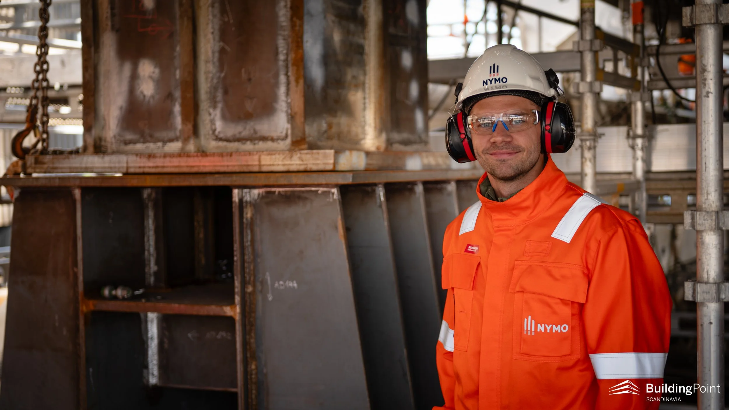 A construction worker wearing safety glasses, ear protection, a white helmet with a NYMO logo, and an orange safety jacket, standing on a construction site with scaffolding and metal beams in the background.