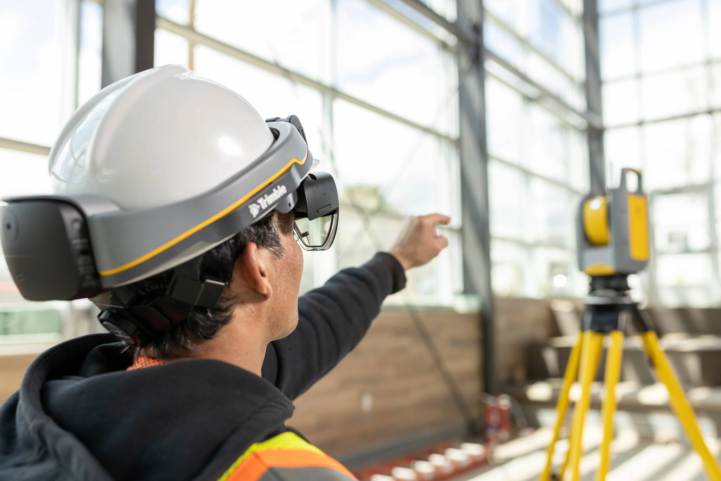 Person wearing a hard hat and augmented reality headset points towards a laser surveying device in a construction site.