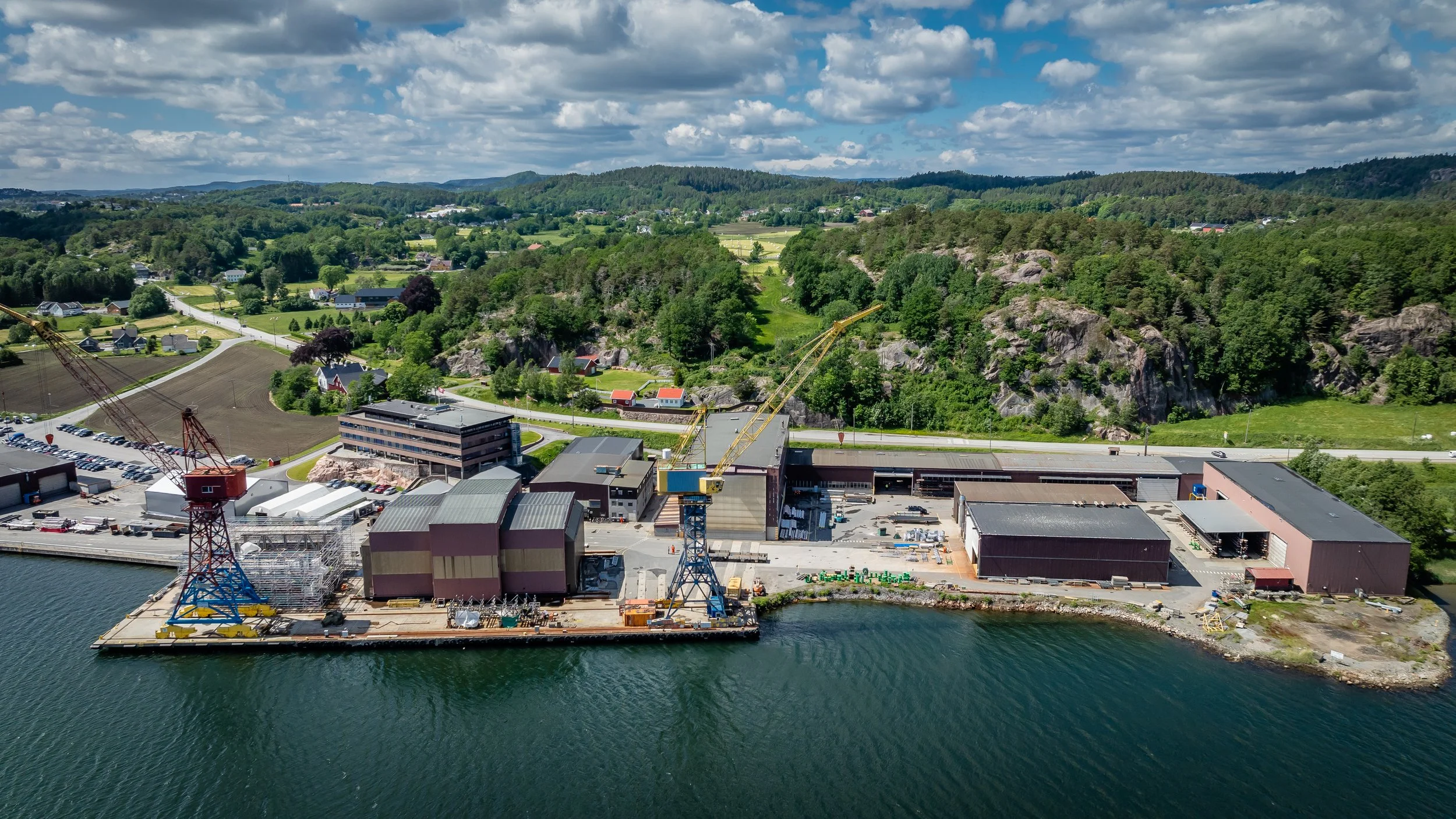 Aerial view of an industrial area near the coastline, surrounded by green hills and trees. Several large buildings and cranes are visible, along with a nearby road and scattered residential houses. The sky is partly cloudy.