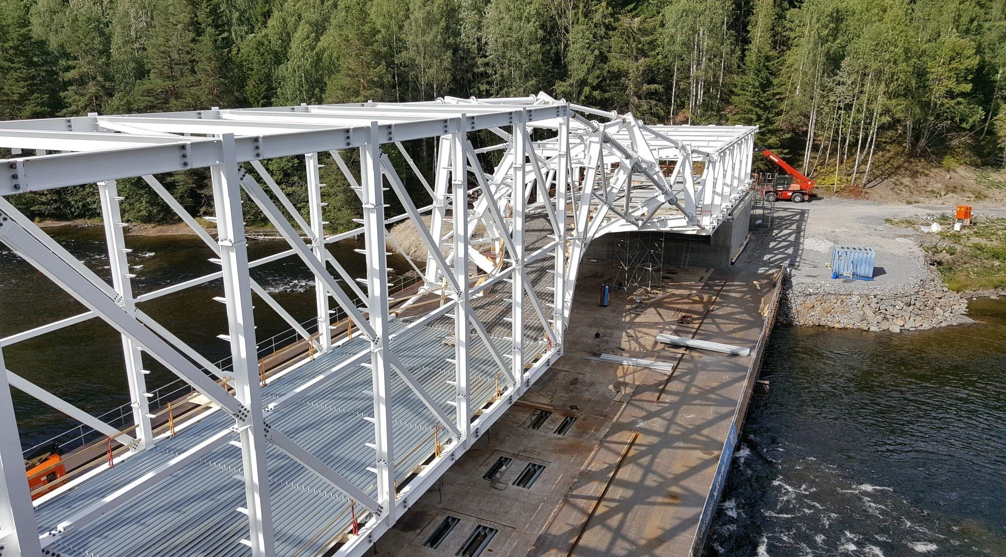 Construction of a steel bridge over a river, surrounded by trees and machinery on a gravel path.