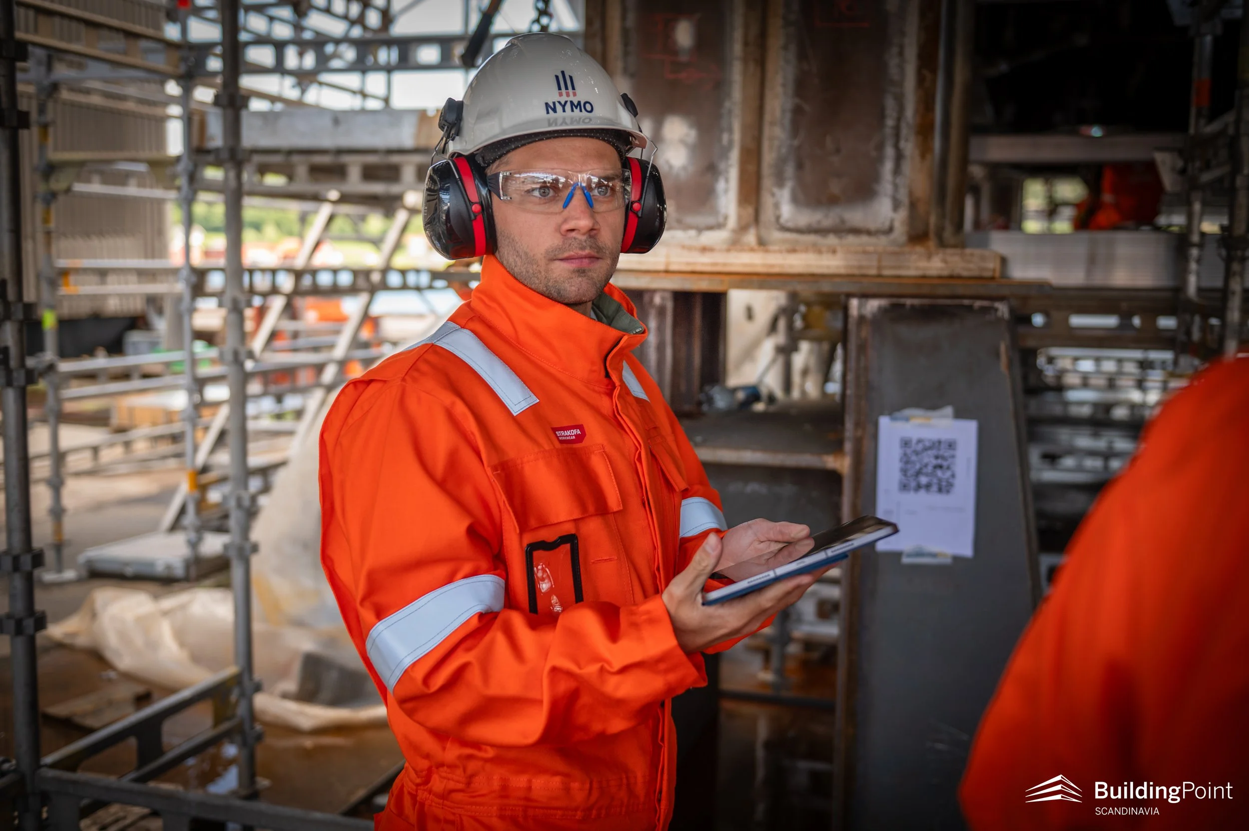 A man wearing a white hard hat, clear safety glasses, orange reflective work jacket, and hearing protection stands inside a construction site holding a tablet. Scaffolding and construction materials are in the background.