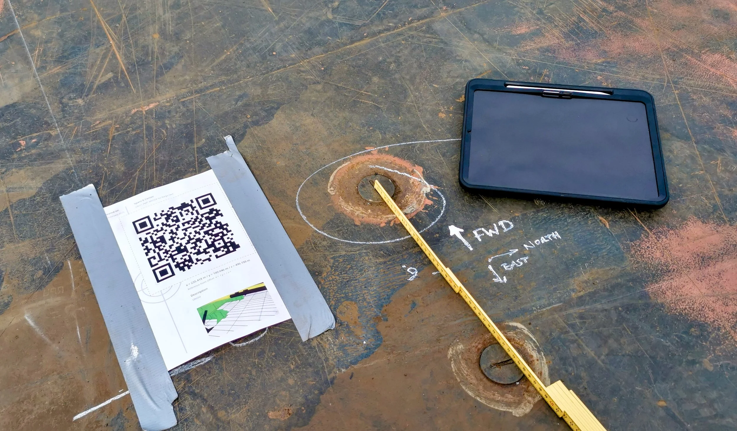 Trimble SiteVision Core - Construction site metal surface with chalk markings, a QR code sheet secured with tape, a digital measuring device, and a yellow measuring tape measuring a rusted circular fixture.