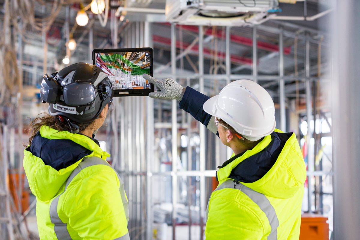 Two construction workers in safety gear, one with a helmet and the other with a hard hat, are reviewing blueprints or plans on a tablet at a construction site with scaffolding.