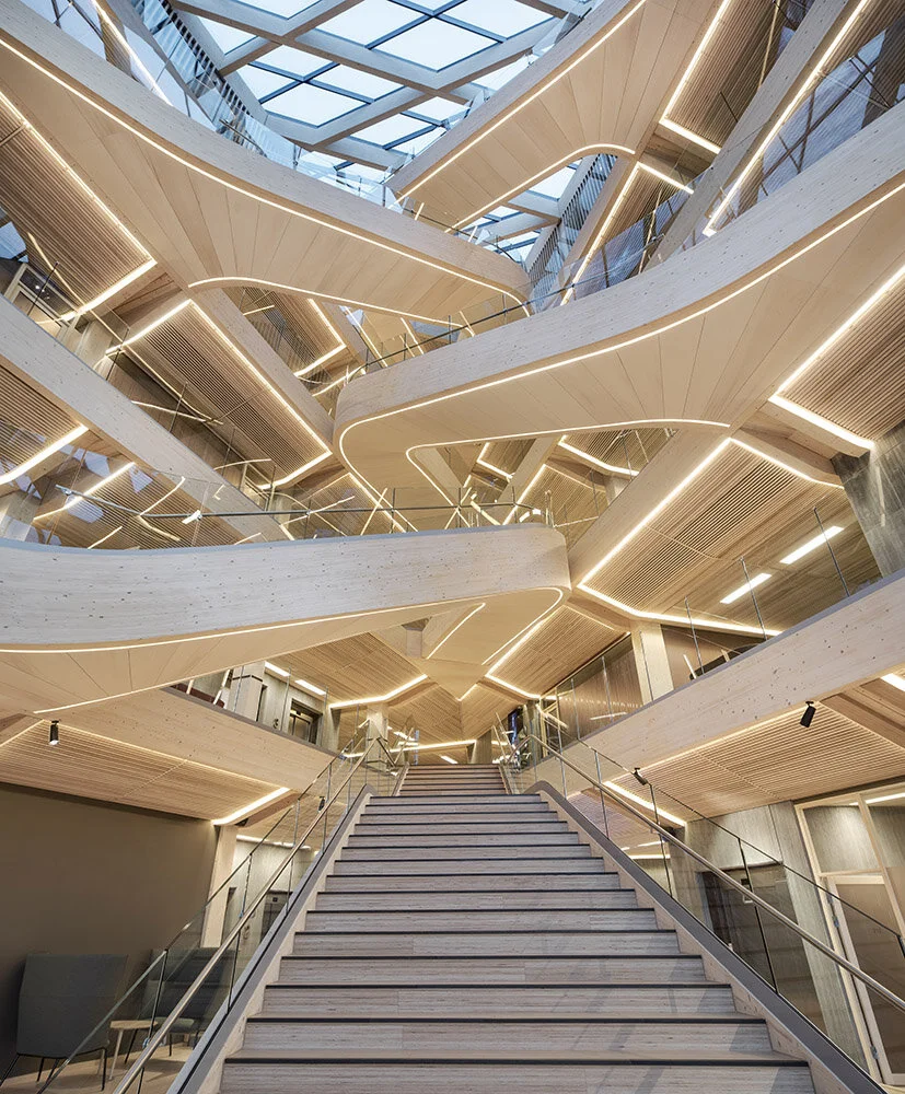 Interior view of a modern building with multiple winding wooden staircases and floors, illuminated by sleek strip lighting, with a glass ceiling at the top.