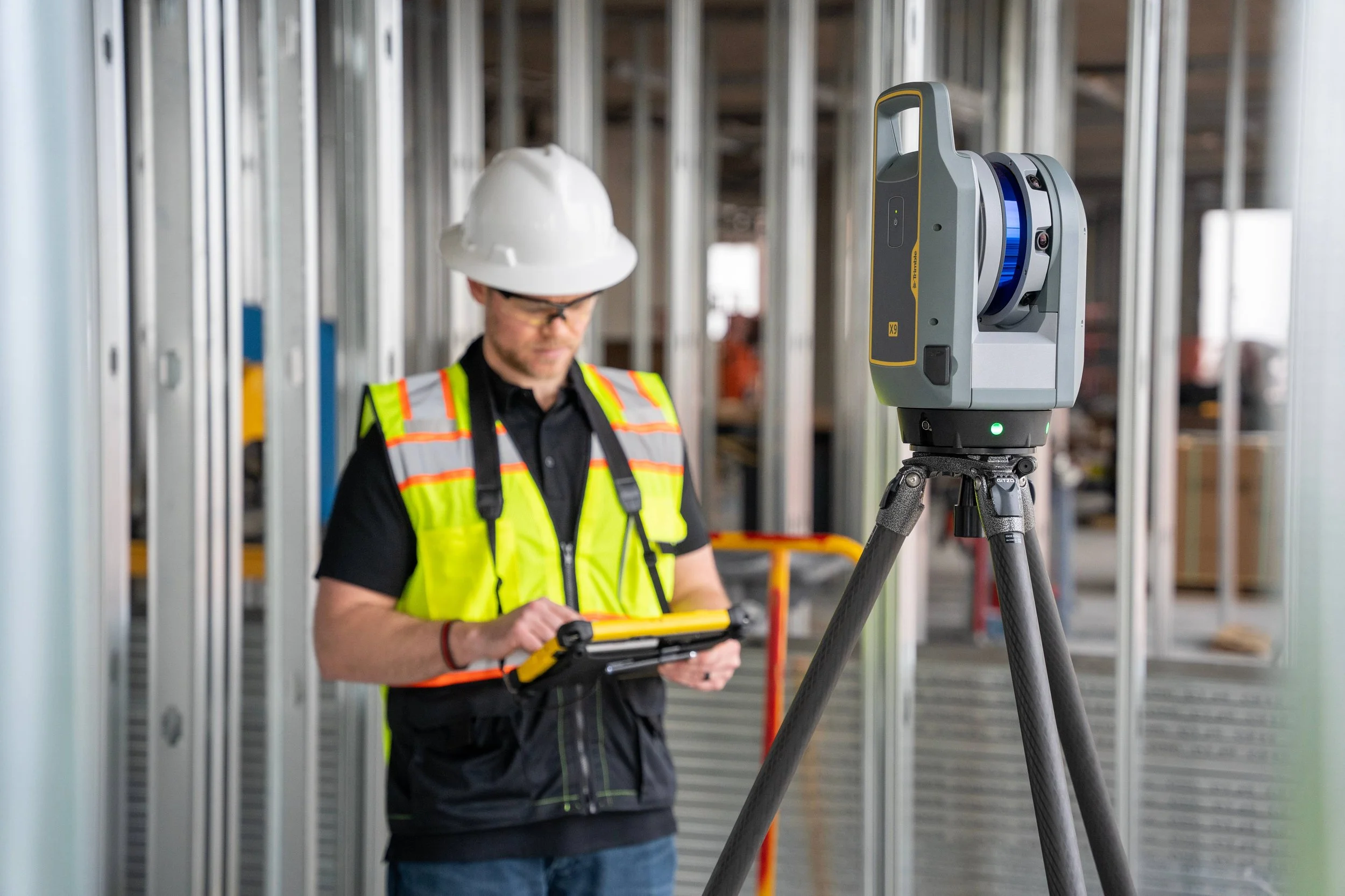 Construction worker in safety gear using a tablet near a surveying instrument on a tripod at a building site.