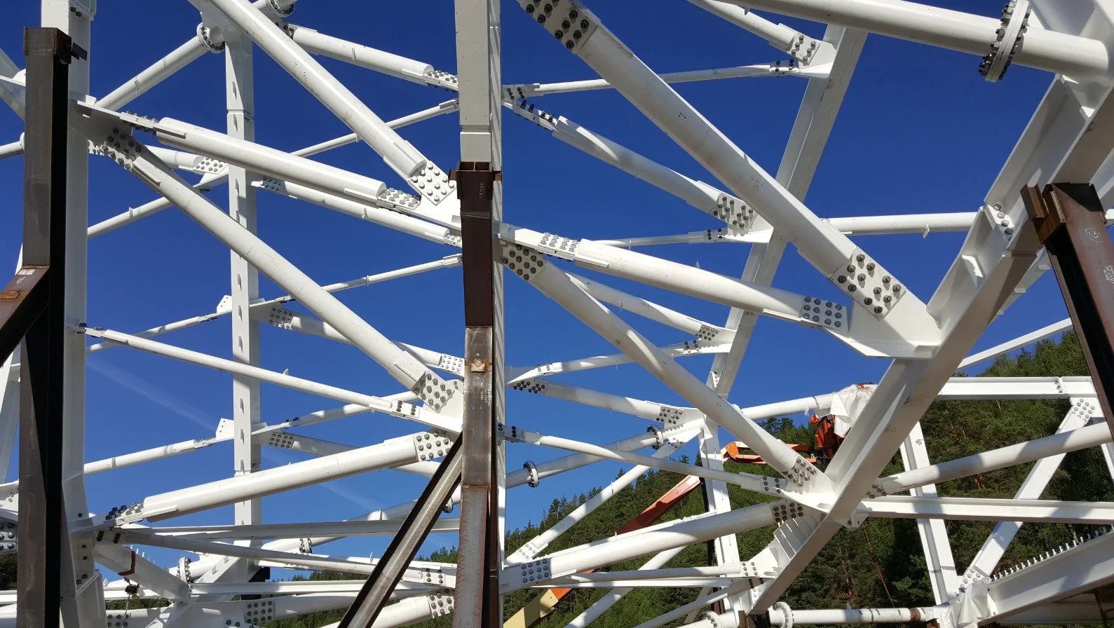Close-up view of a large white metal structure with interconnected beams and bolts, set against a clear blue sky and a hint of green trees in the background.