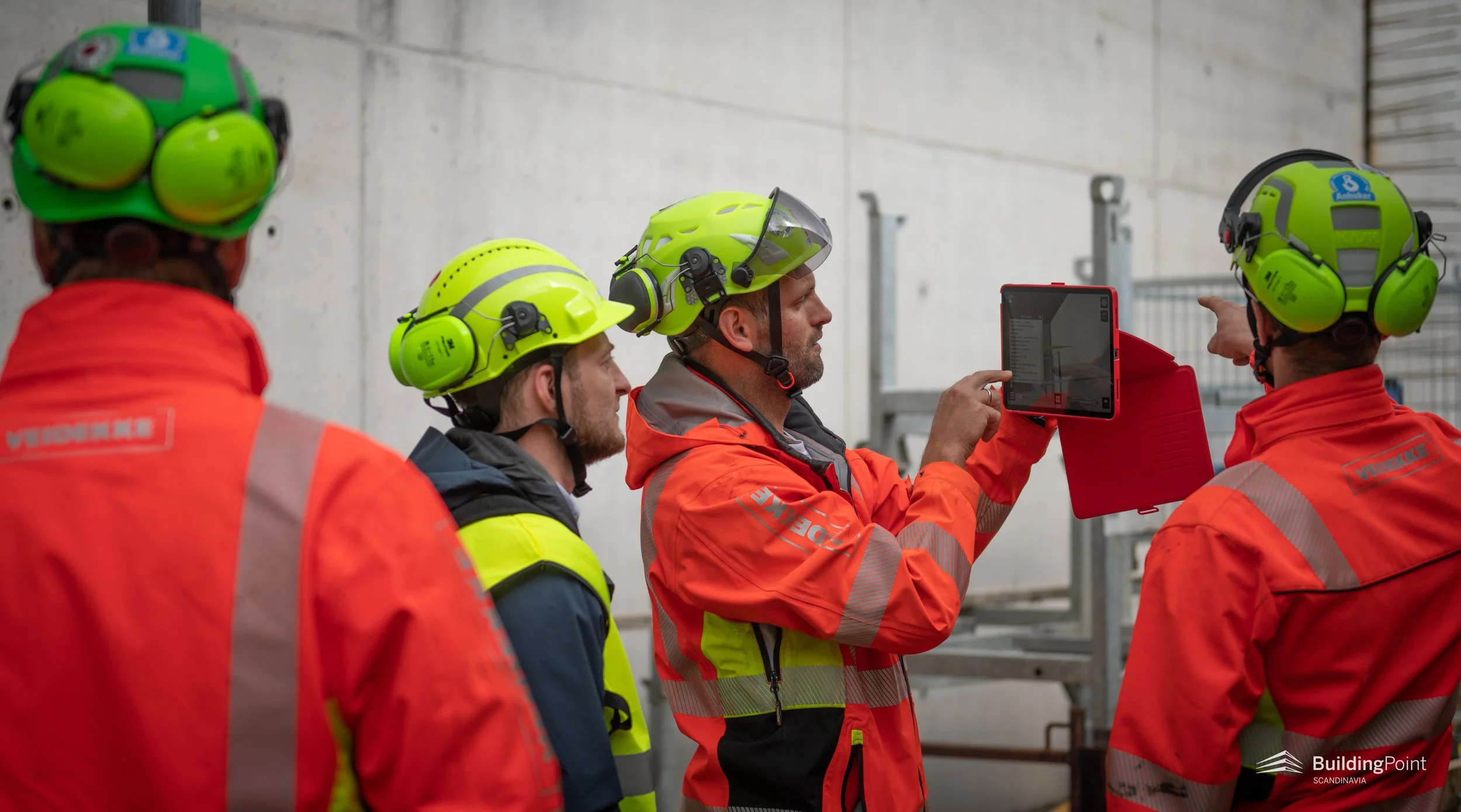 Group of construction workers wearing high-visibility jackets and safety helmets, gathered around a supervisor who is holding a tablet device and explaining something. The workers are attentively listening, with a concrete wall and scaffolding in the background.