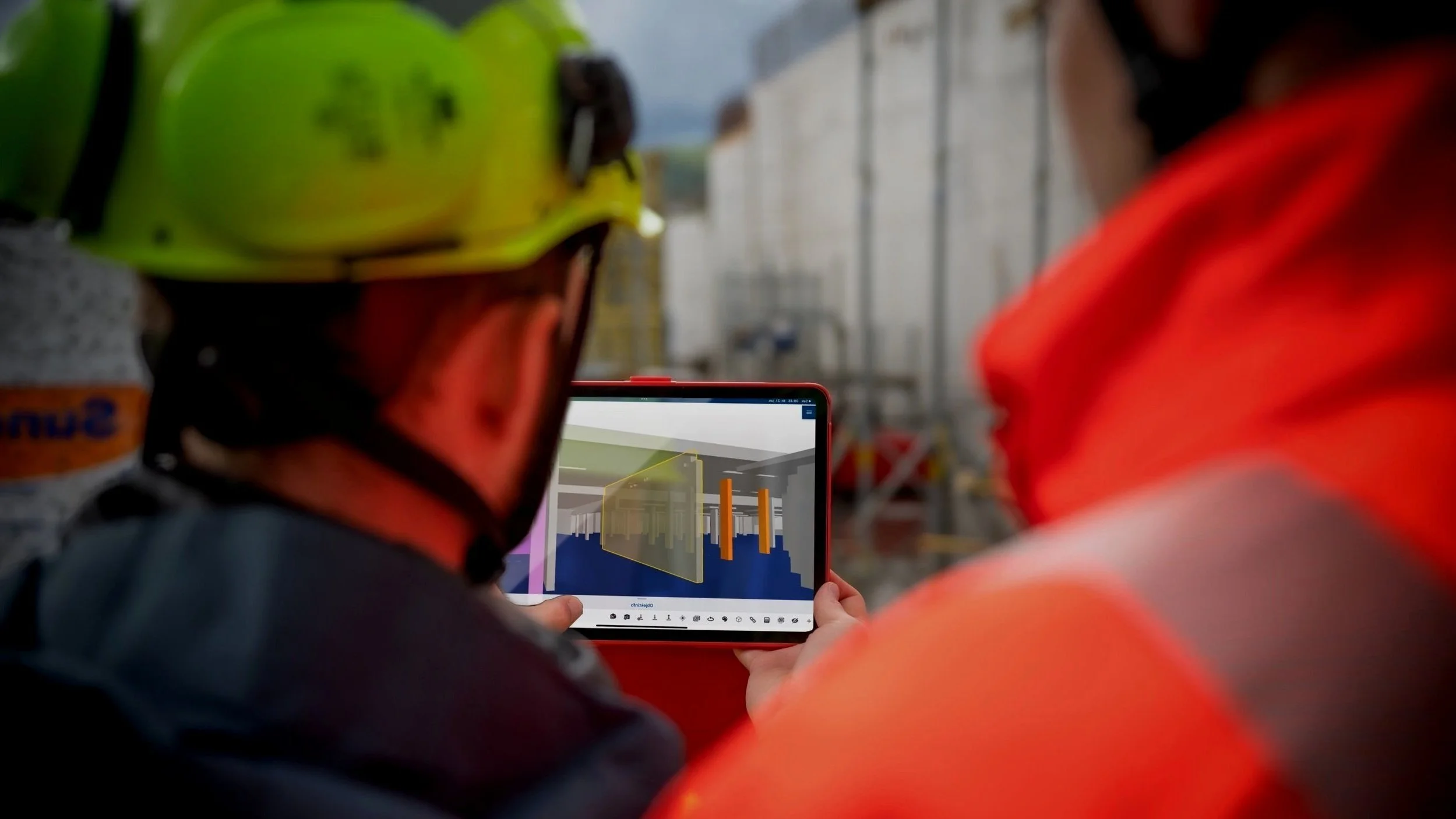 Two construction workers wearing helmets and safety vests look at a tablet displaying a digital 3D model of an underground construction project.