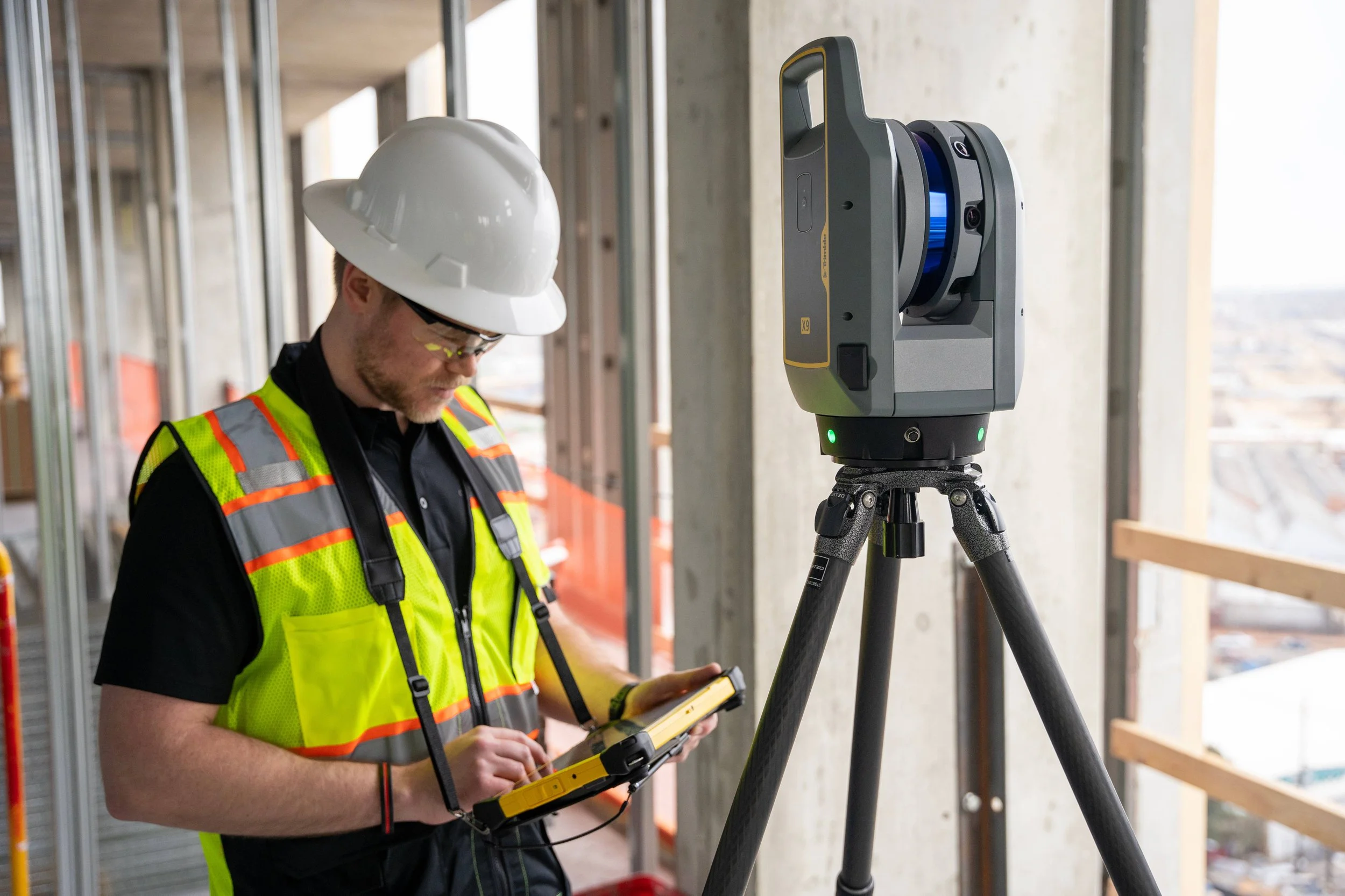 A construction worker wearing a white hard hat and a neon yellow safety vest operating a surveying instrument on a tripod at a building site.
