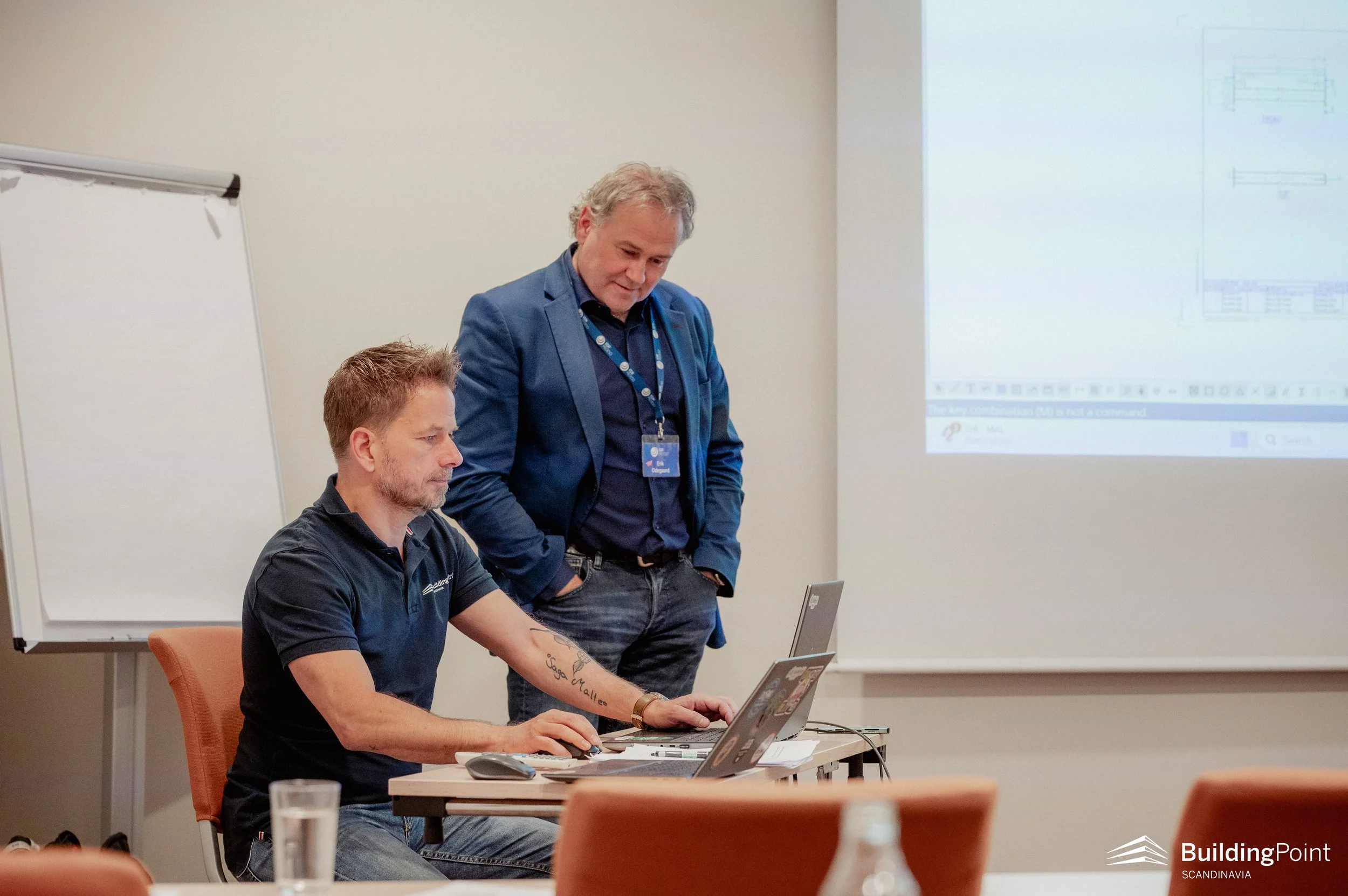 Two men are in a meeting room; one is seated at a desk working on a laptop, and the other stands beside him looking at the screen. The seated man has tattoos on his arm, and the standing man is dressed in a blue blazer and looks intently at the laptop. There is a white projector screen in the background showing a presentation, and a whiteboard on the side.