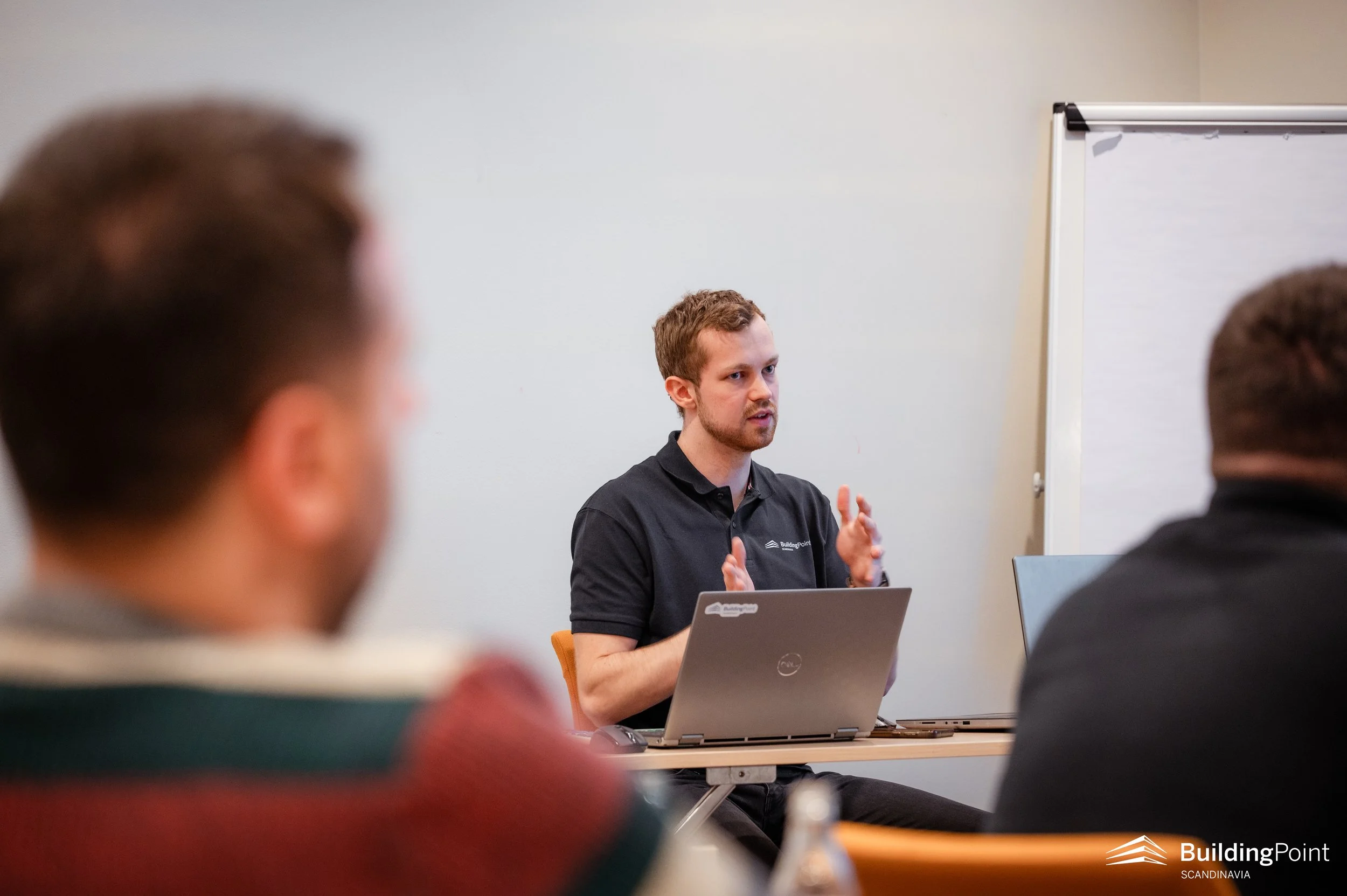 A man in a black polo shirt is speaking and gesturing with his hands during a presentation in a conference room. He is seated at a table with a laptop in front of him, and there are other people in the audience. A whiteboard is visible in the background.