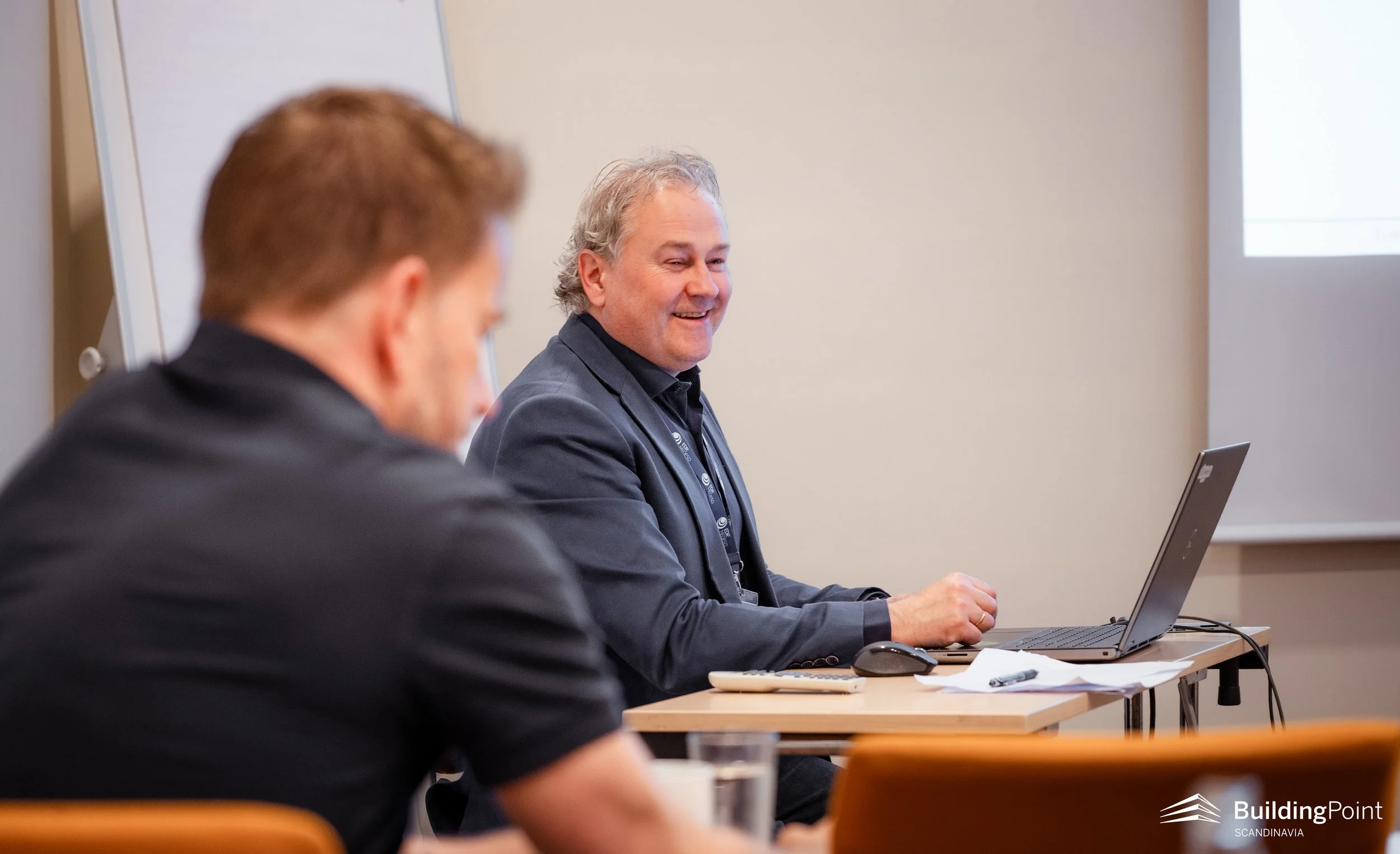 A man with gray hair and a dark gray shirt smiling while sitting at a conference table, with a laptop in front of him and another person with reddish-brown hair blurred in the foreground. The setting appears to be a meeting or seminar room with a whiteboard behind them.