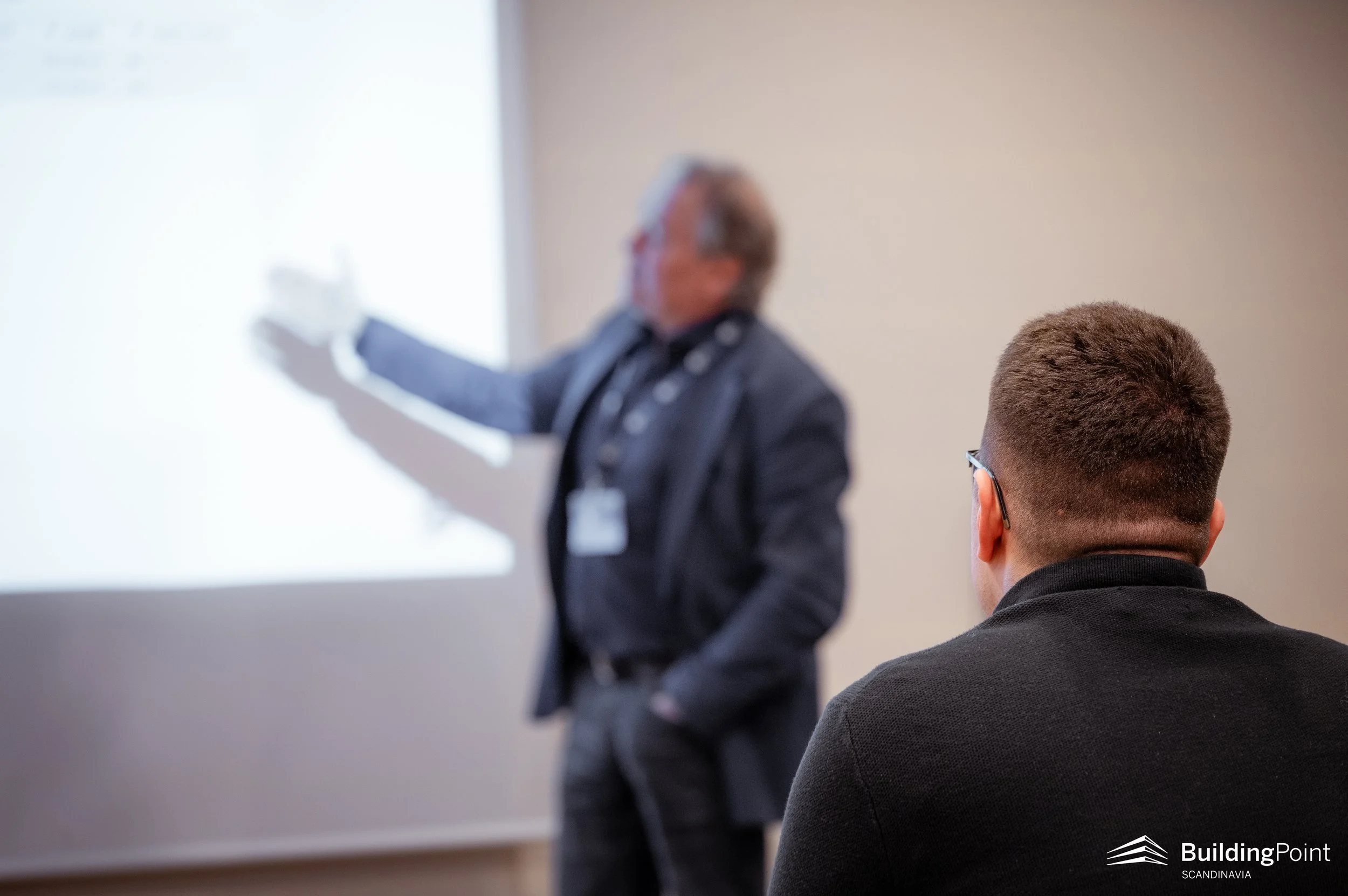 A person with short hair watches a presentation while a male presenter, dressed in a dark suit, points at a screen with a white background. The photo is taken indoors during a professional or educational event.