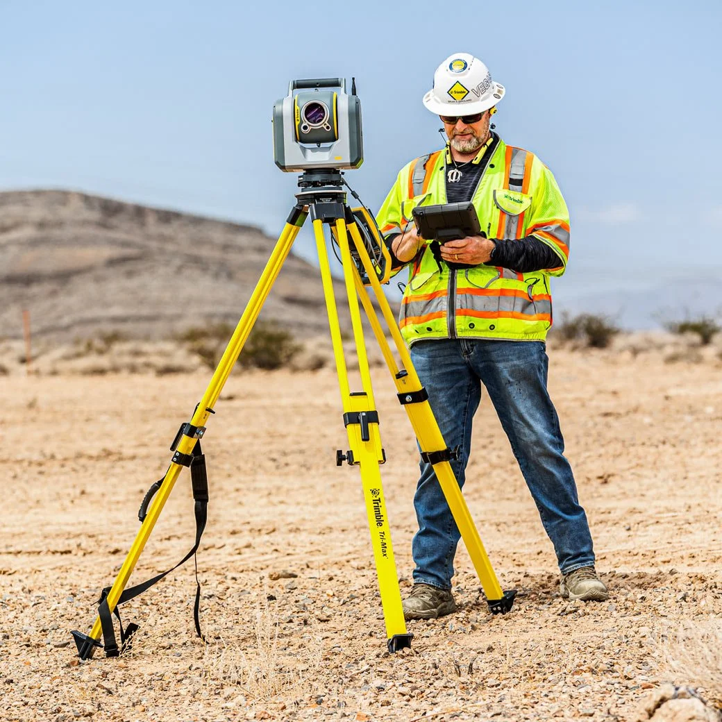 A surveyor in a yellow safety vest and hard hat stands outdoors in a desert-like area, using a tablet with a yellow tripod-mounted surveying instrument.