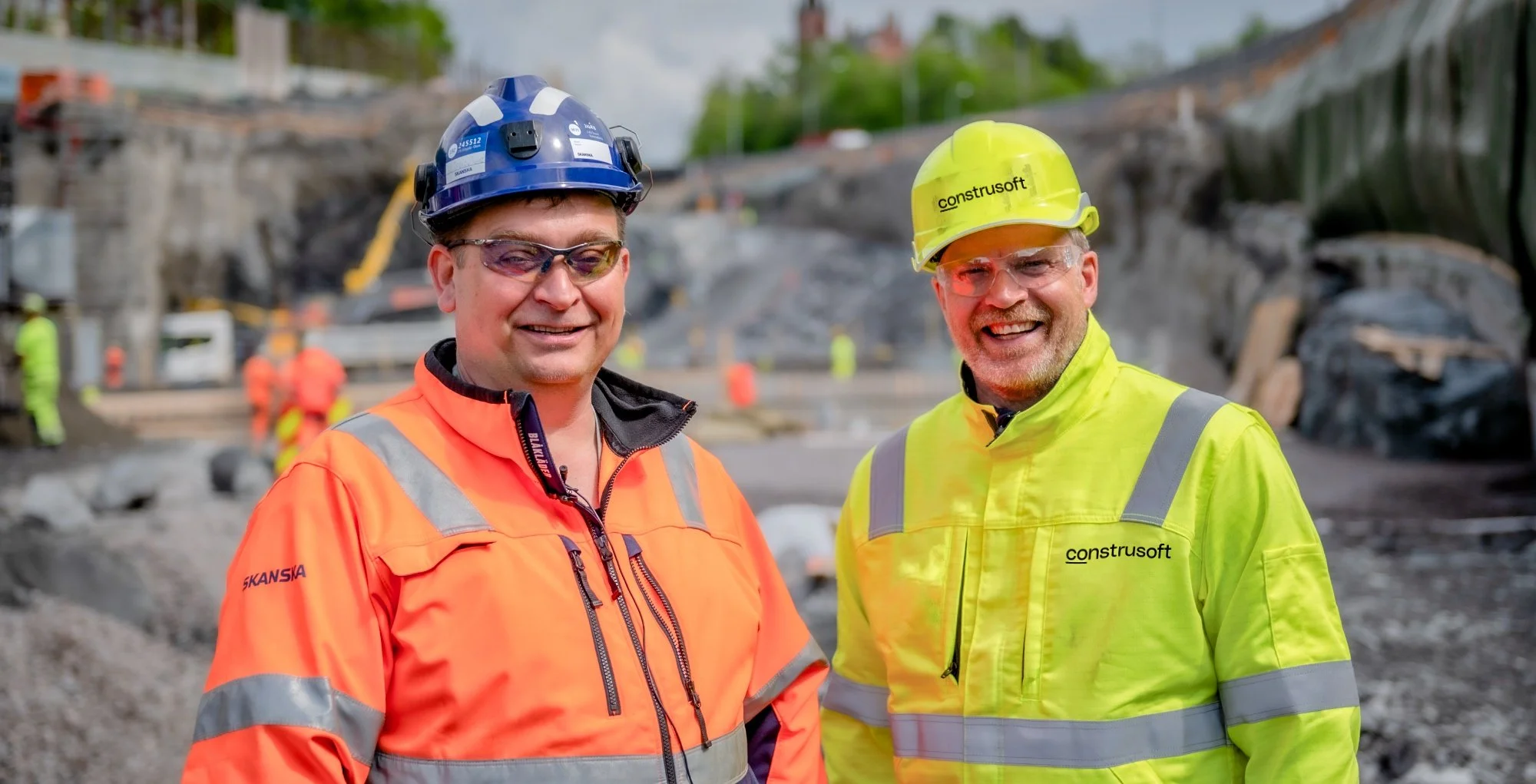 Anders from Construsoft and Bård Olav in Skanska on a construction site, both wearing high-visibility jackets and hard hats, with equipment and workers in the background.