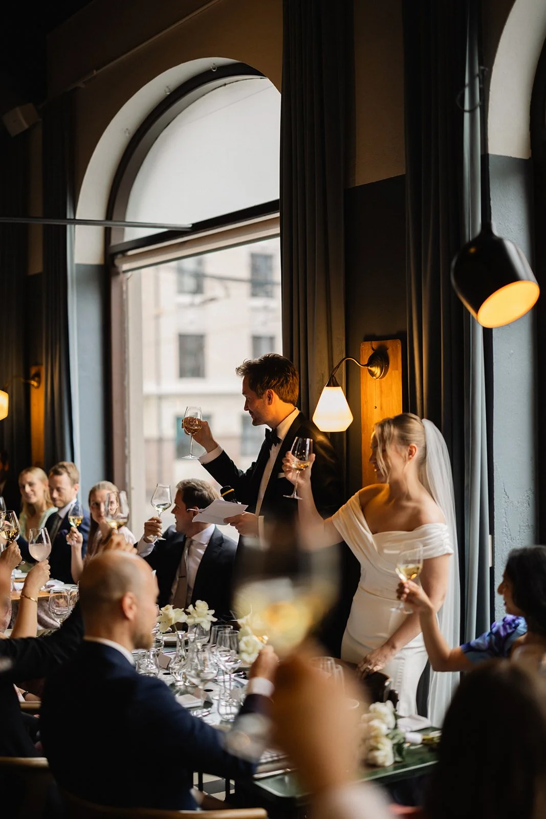 Brudepar avholder en tale under et bryllupsfeiring med gjester som holder glassene oppe.