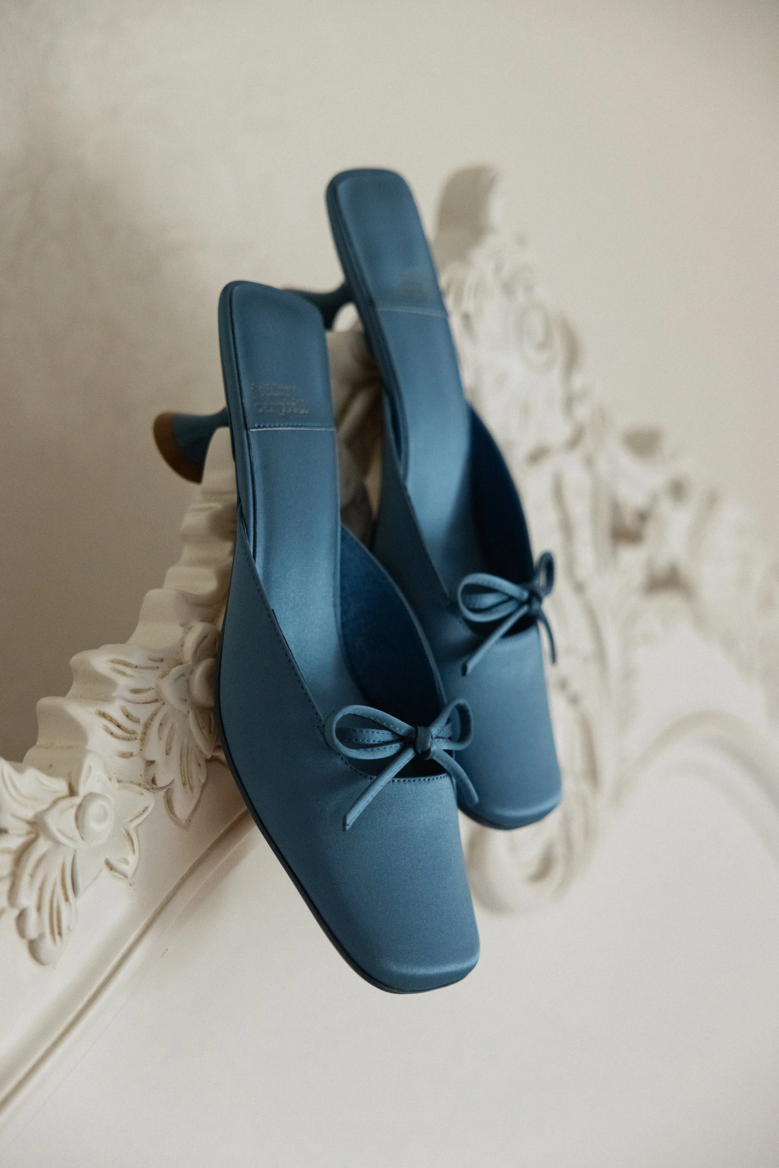 Et par blå sko med hæler og sløyfer på tåen, plassert på en ornamentert hvit overflate.